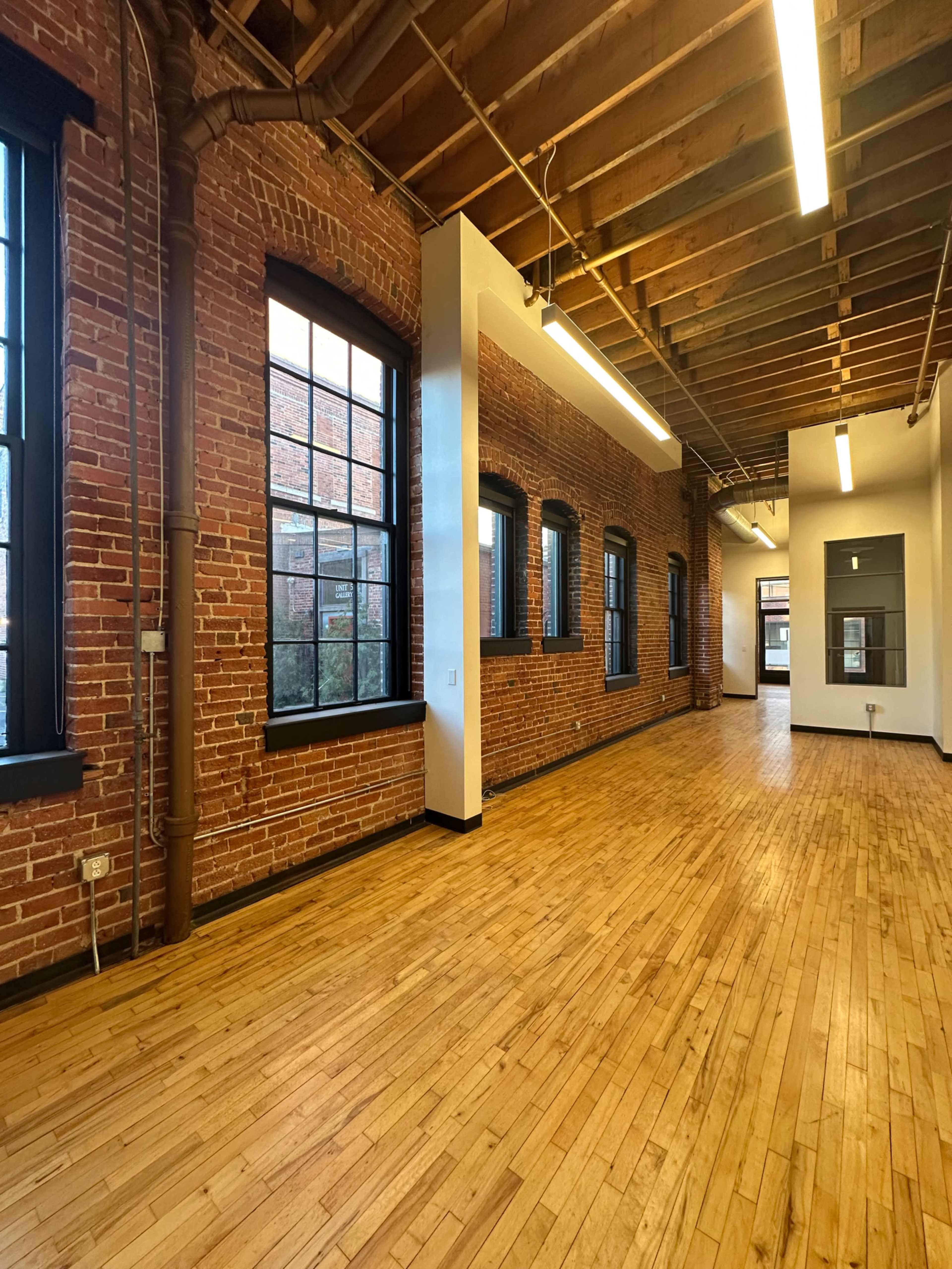 The image shows an empty room with exposed brick walls, large windows, and hardwood floors, featuring a high ceiling with visible beams.