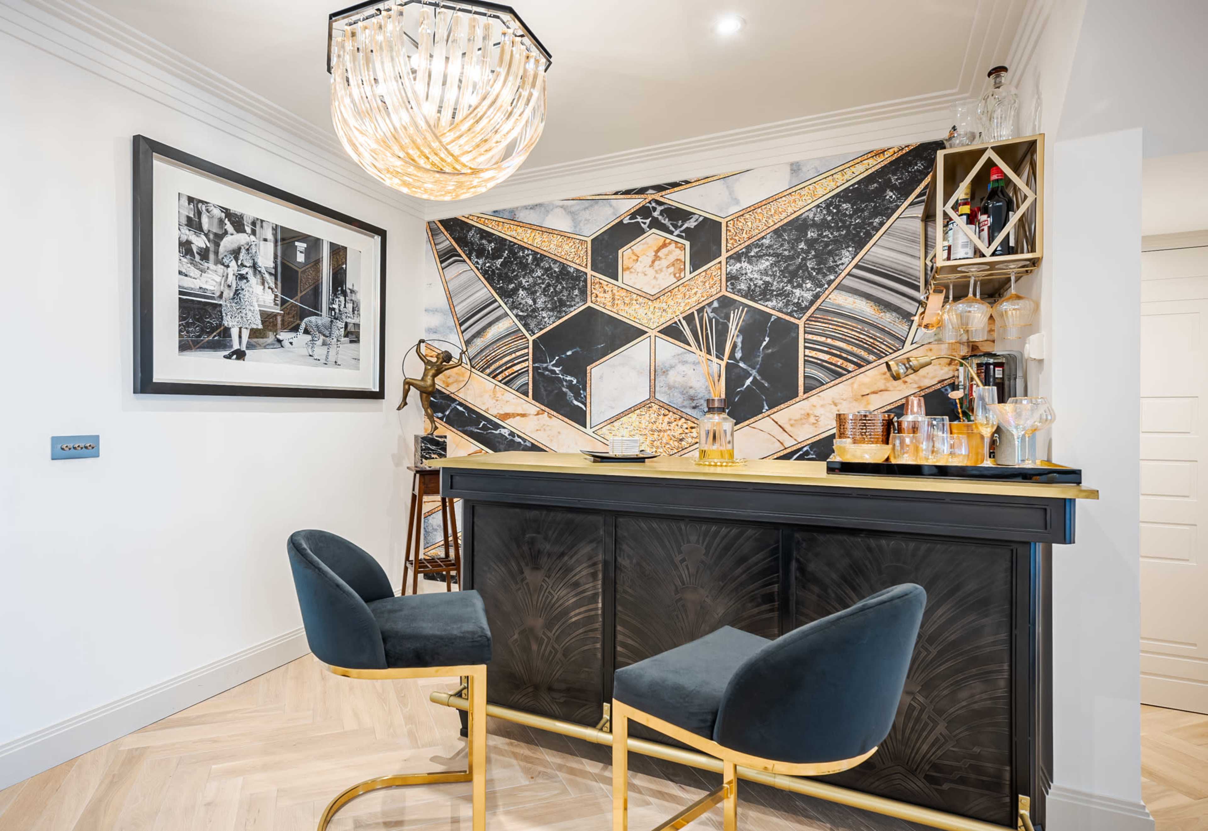 The image shows a modern home bar with a geometric patterned wall, a dark wooden bar counter, and two velvet stools.