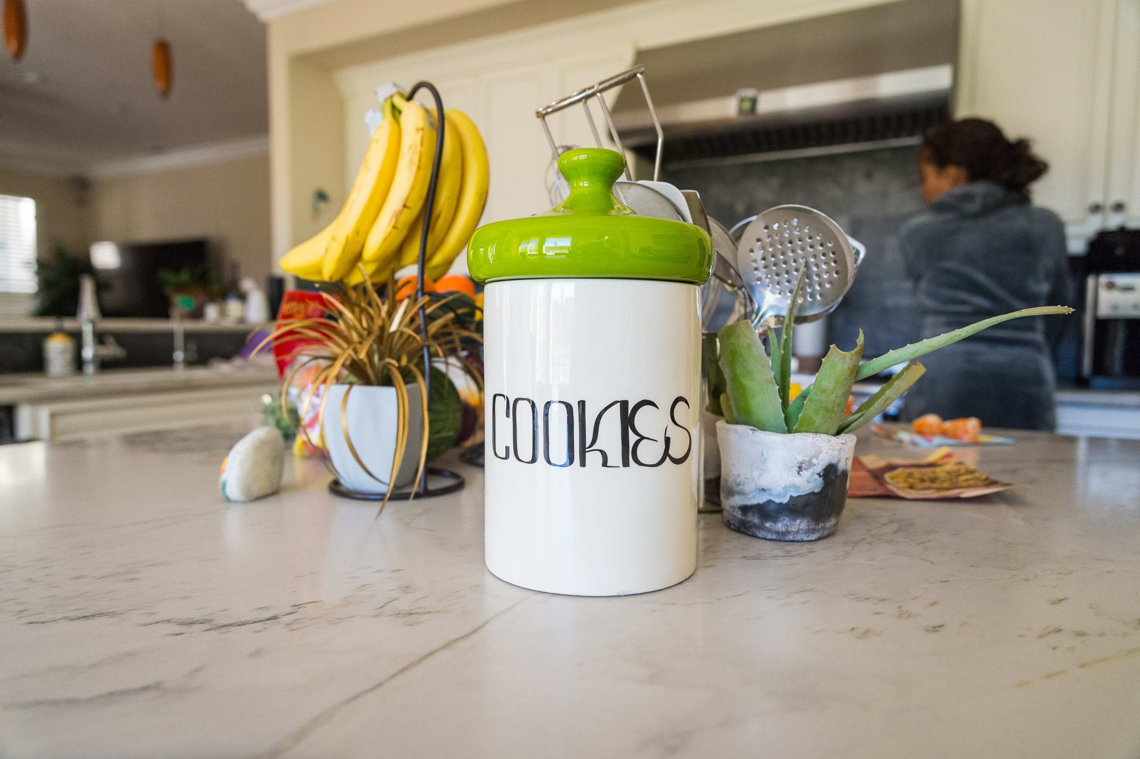 A white cookie jar with a green lid sits on a marble kitchen countertop, surrounded by plants, bananas, and kitchen utensils.