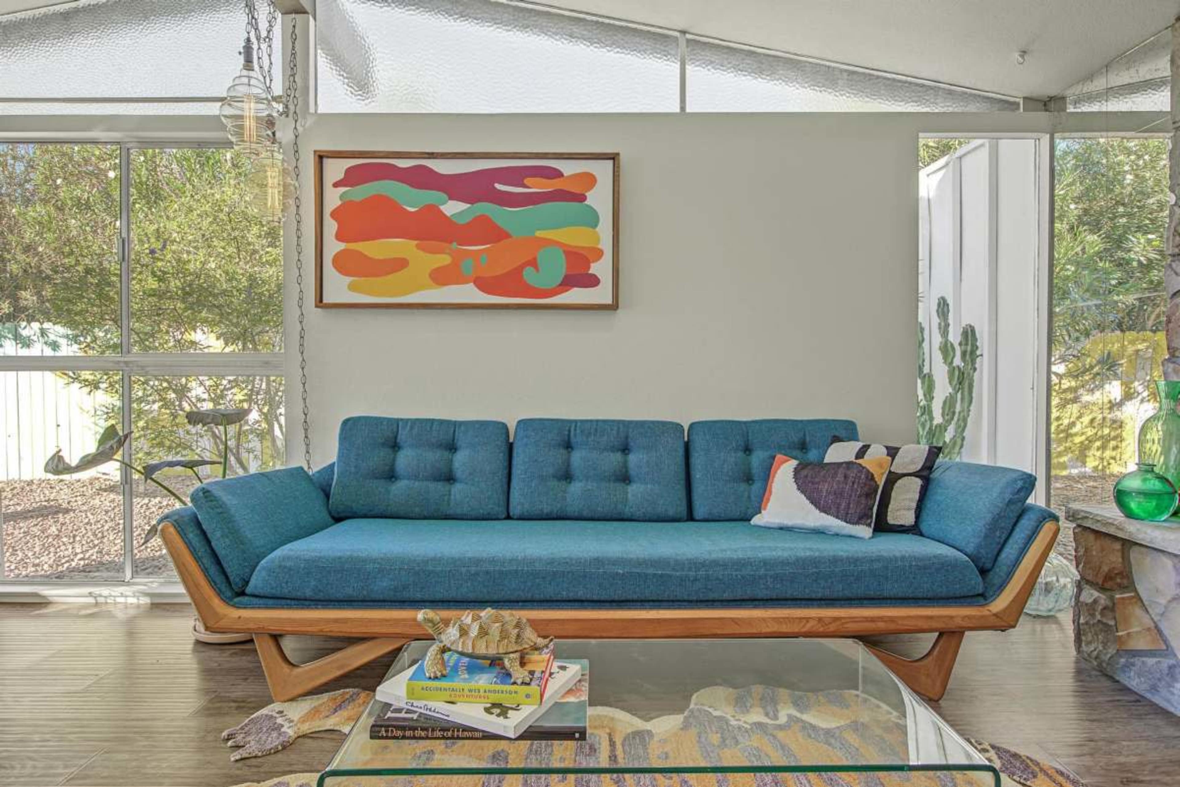 The image shows a modern living room featuring a blue sofa with wooden legs, a colorful abstract artwork on the wall, and a glass coffee table surrounded by decorative items.
