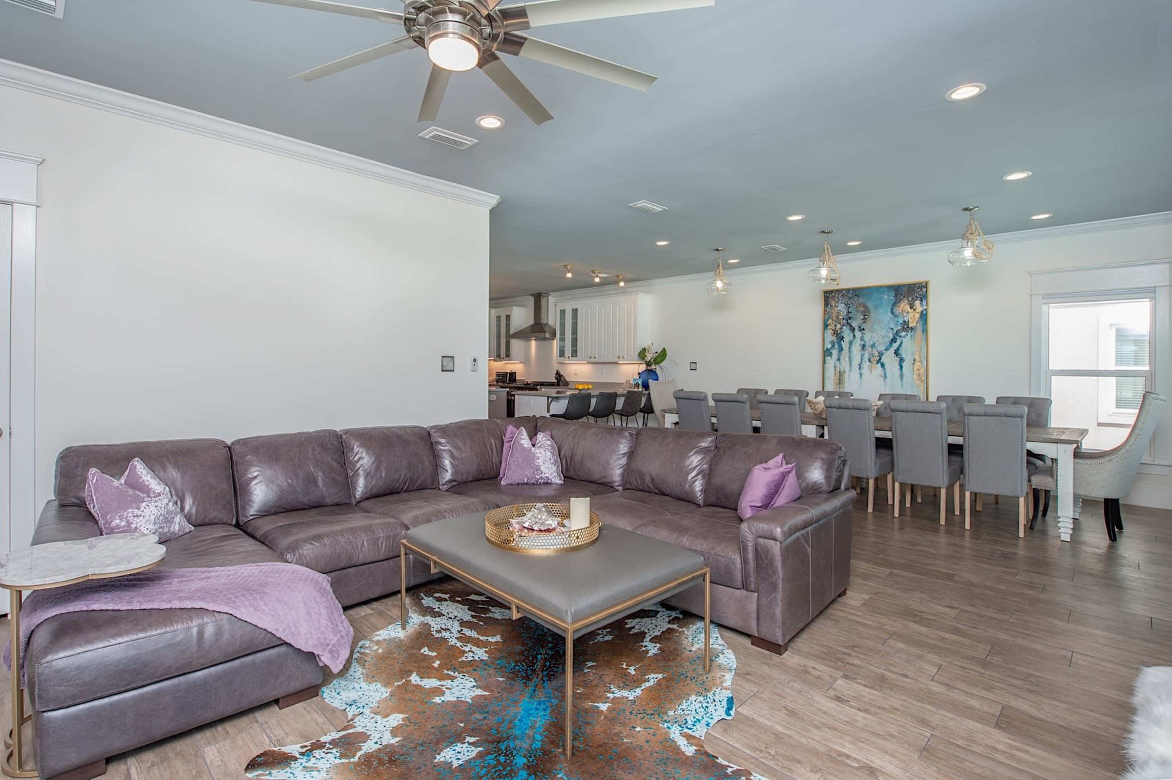 A modern living room with a large sectional sofa, a coffee table, and a dining area in the background featuring a long table and decorative art.