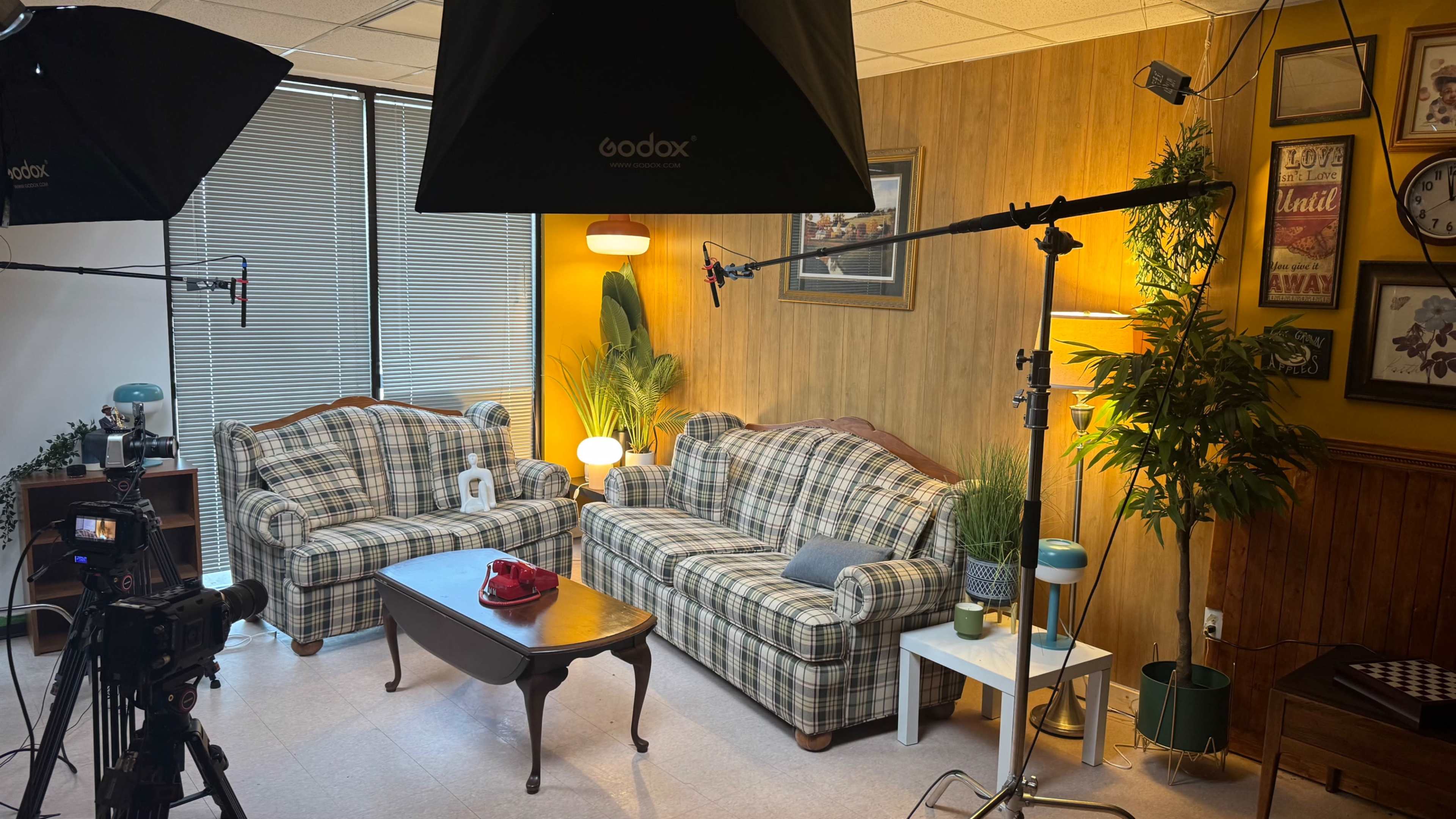 The image shows a cozy indoor setting with two plaid sofas, a coffee table, and studio lighting equipment arranged for filming.