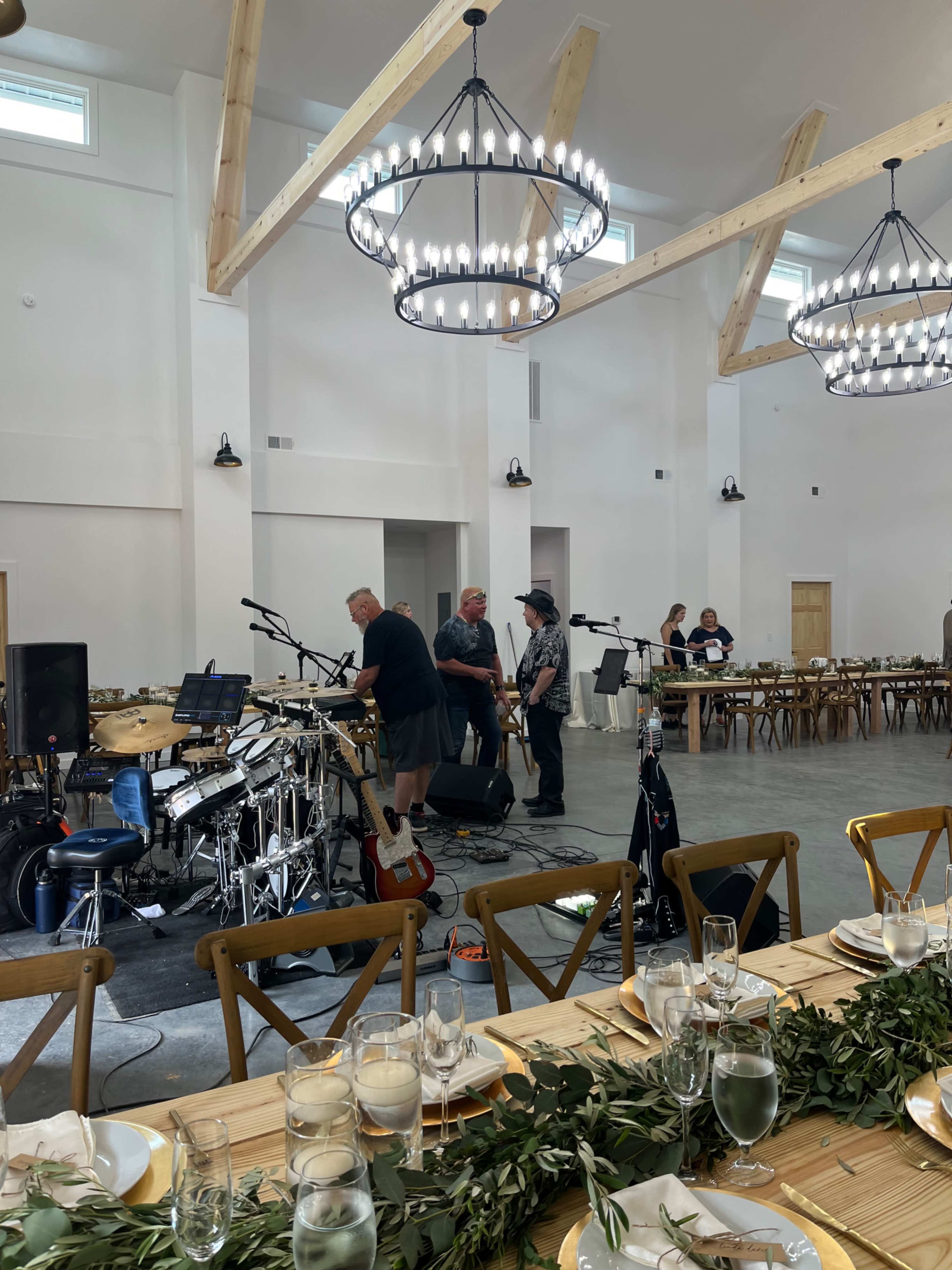A band is setting up their equipment on a raised stage in a spacious event hall with long wooden tables and decorative chandeliers.