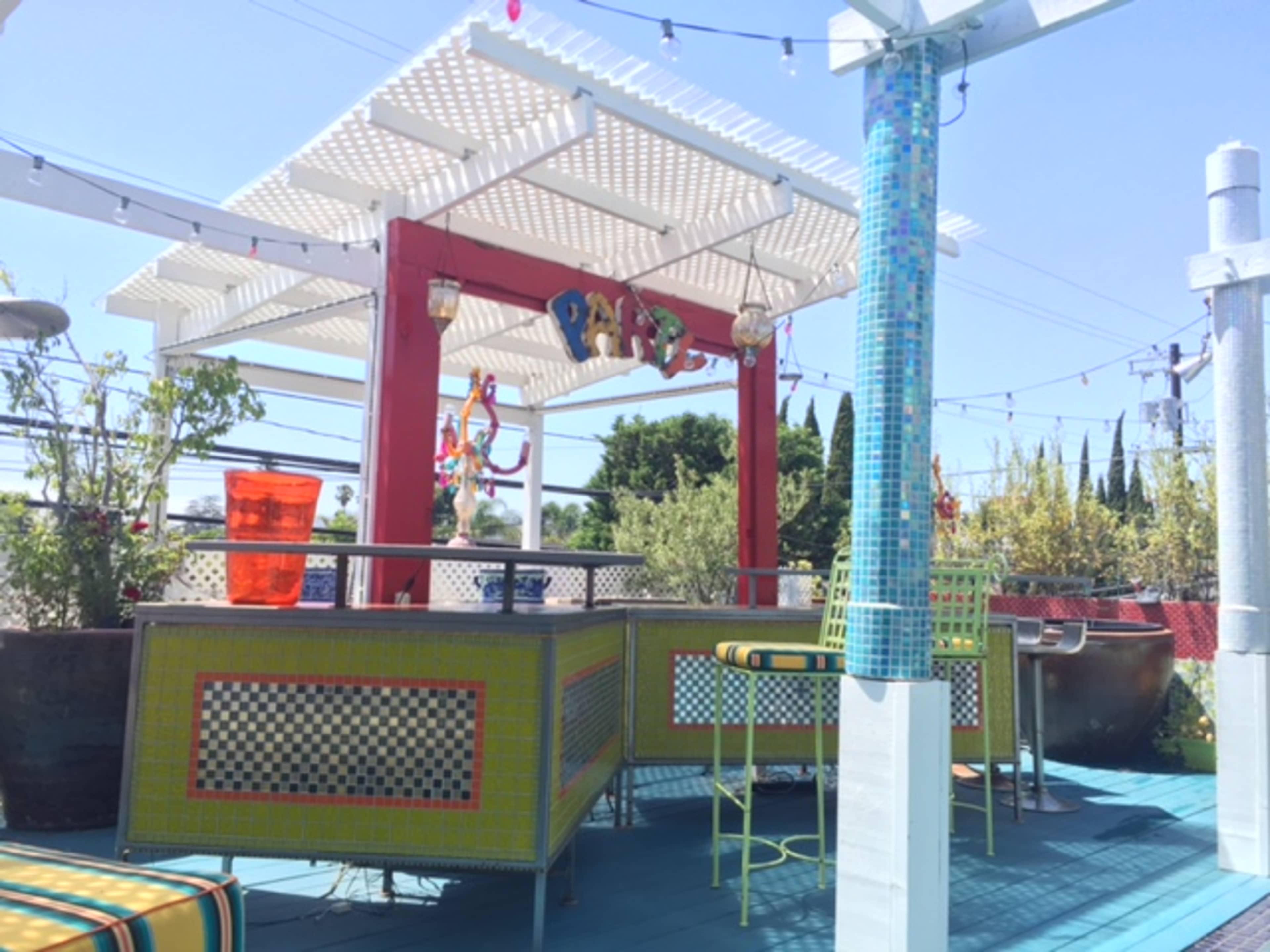 A colorful outdoor bar area features a large shaded pergola with decorative elements, surrounded by greenery and bright furniture.