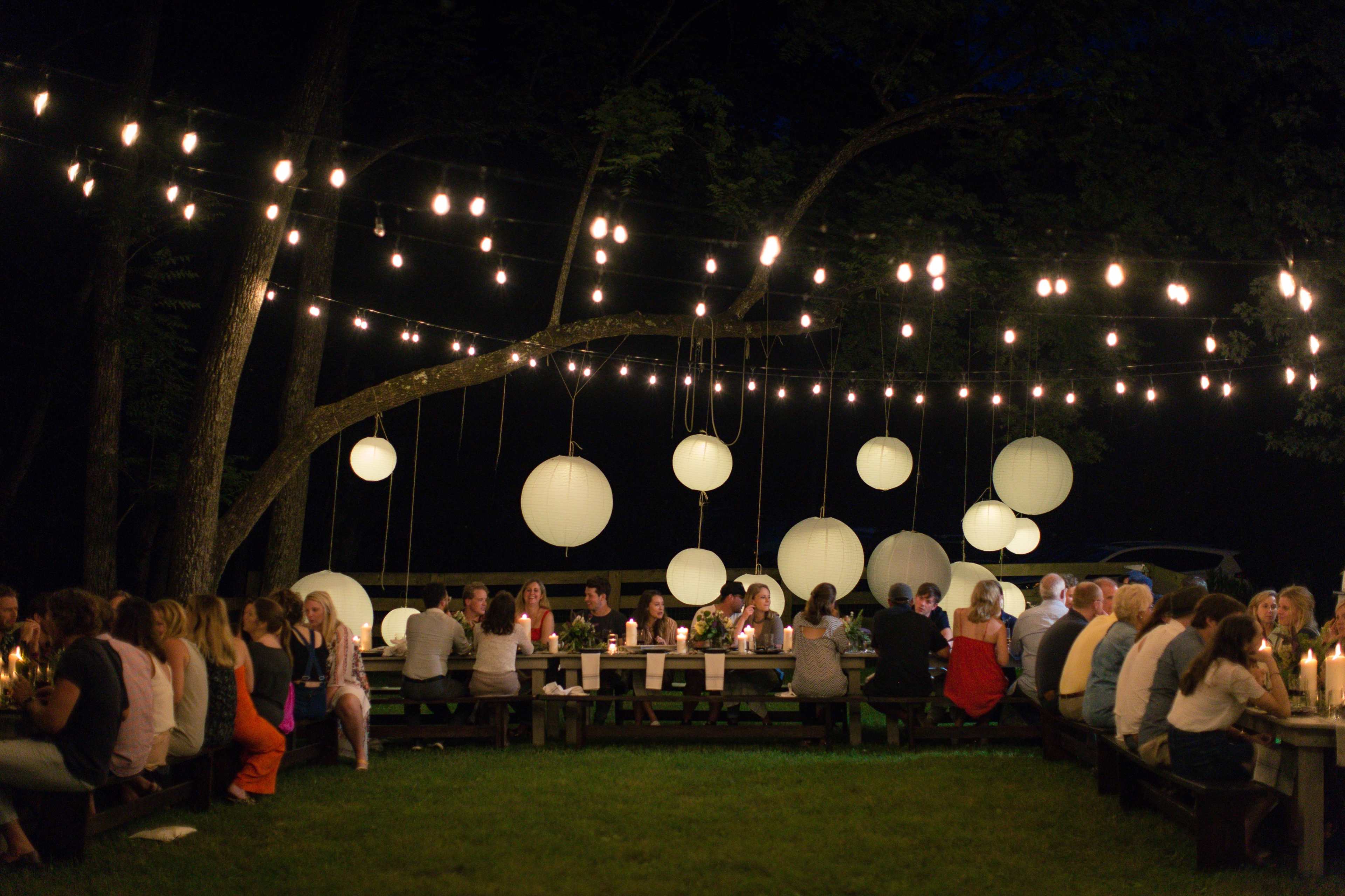 A large gathering of people sits at long tables outdoors, illuminated by string lights and lanterns hanging from trees.