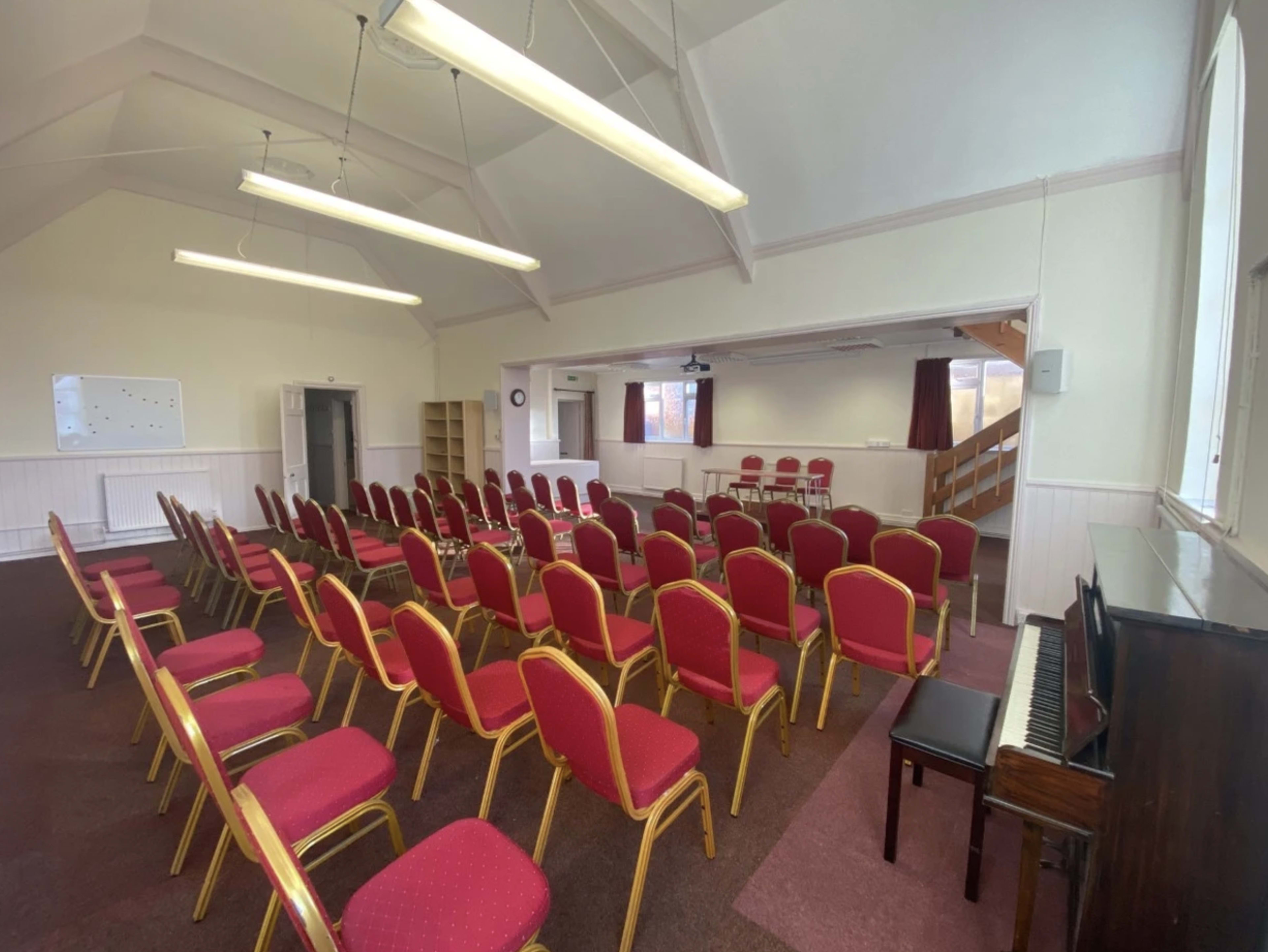 An interior room set up for a gathering, with rows of red chairs facing a small stage, a piano in the corner, and a whiteboard on the wall.