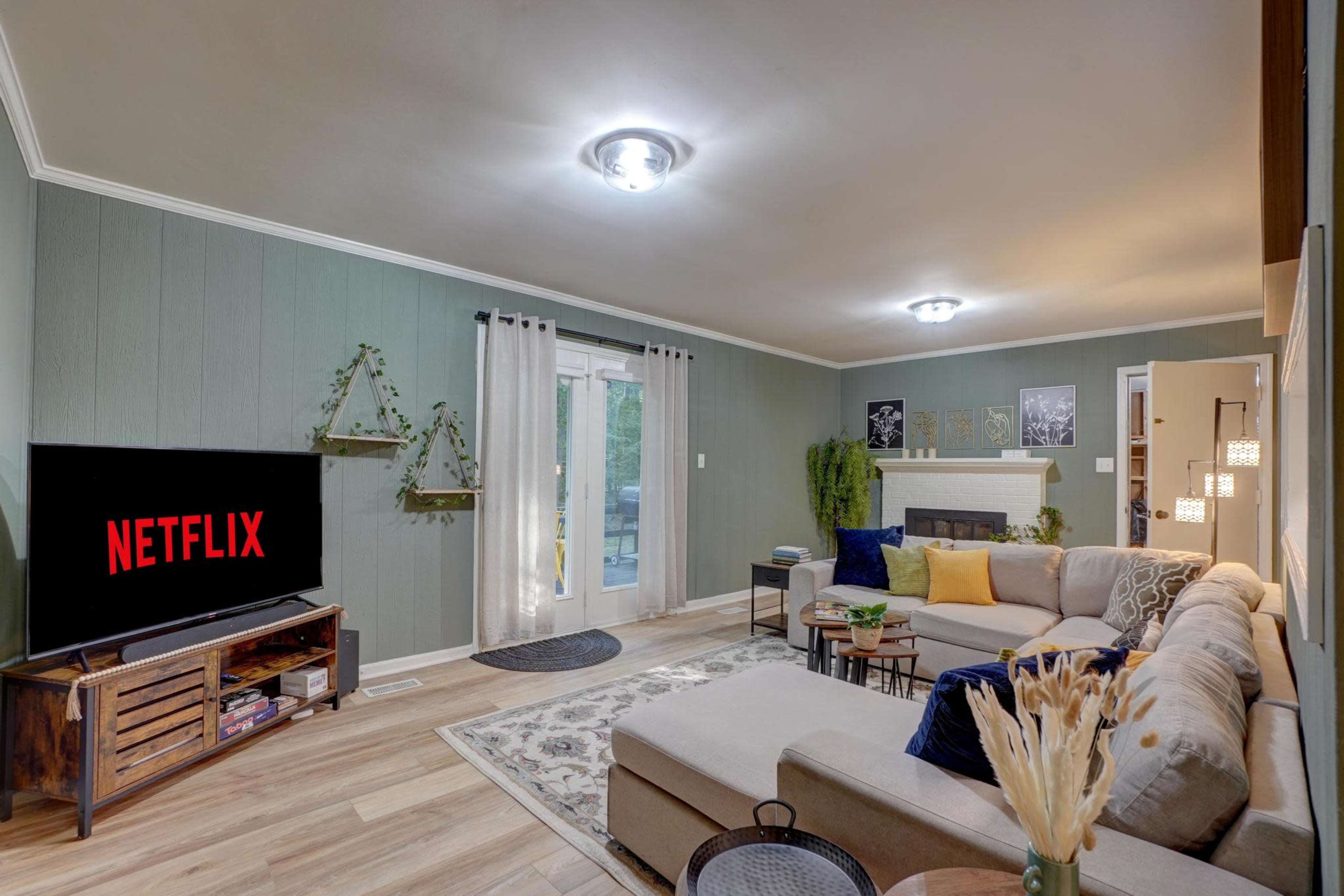 The living room features a sectional sofa, a television displaying the Netflix logo, and decorative plants on the walls.
