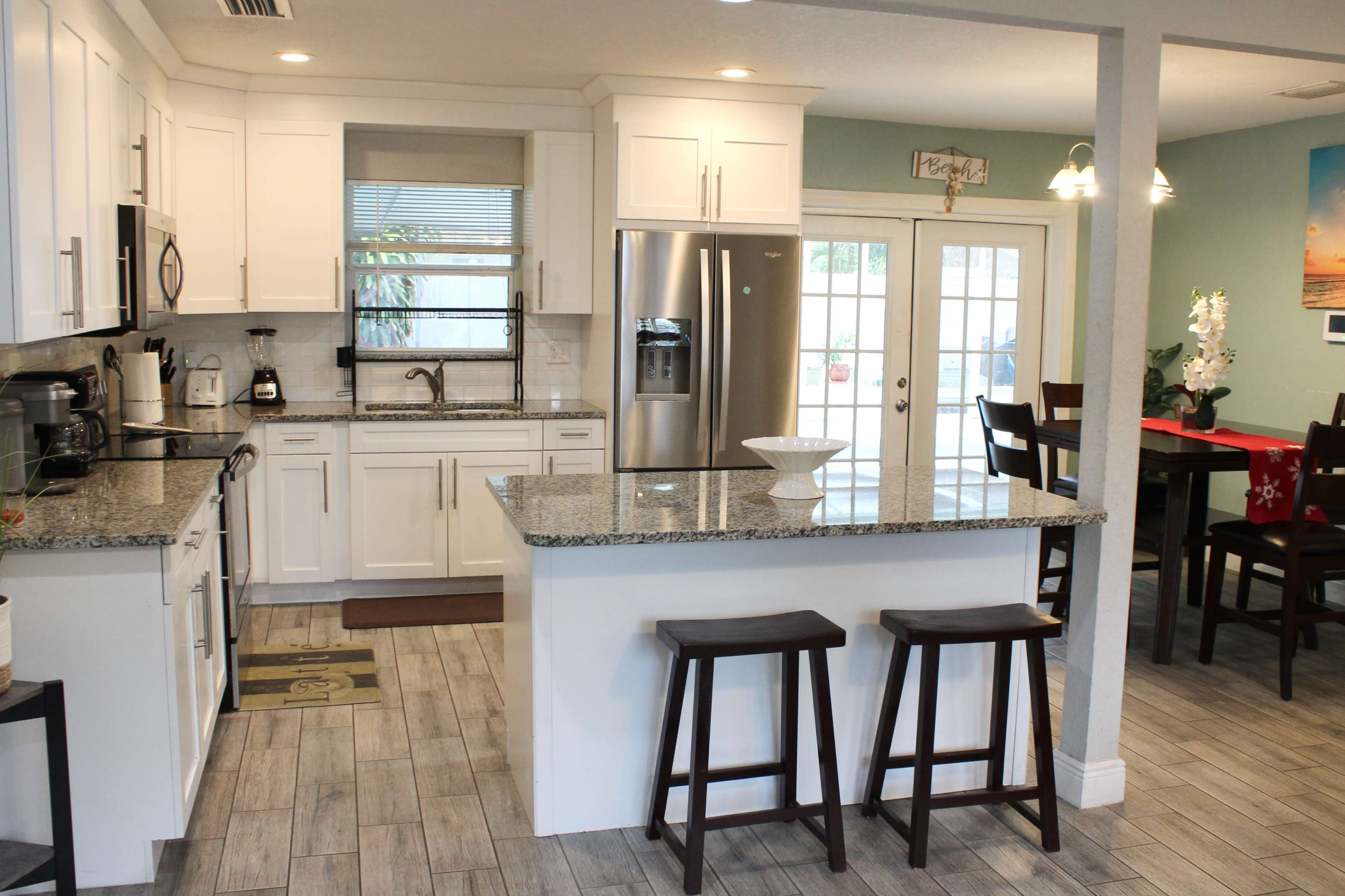 A modern kitchen featuring white cabinetry, granite countertops, and a breakfast bar with two stools, adjacent to a dining area with a table and chairs.
