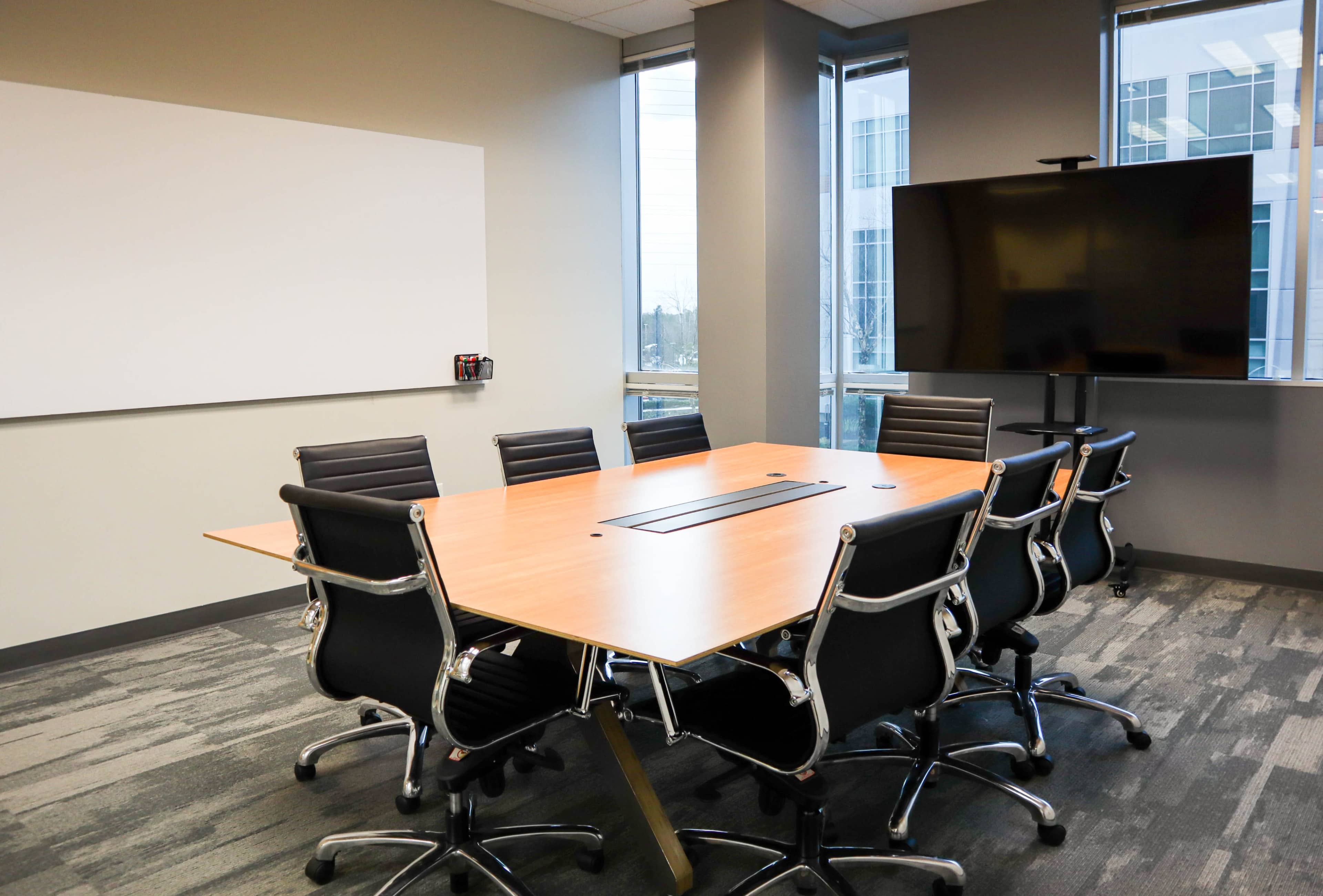 A modern conference room features a large wooden table surrounded by black office chairs, with a whiteboard and a large screen on opposite walls.