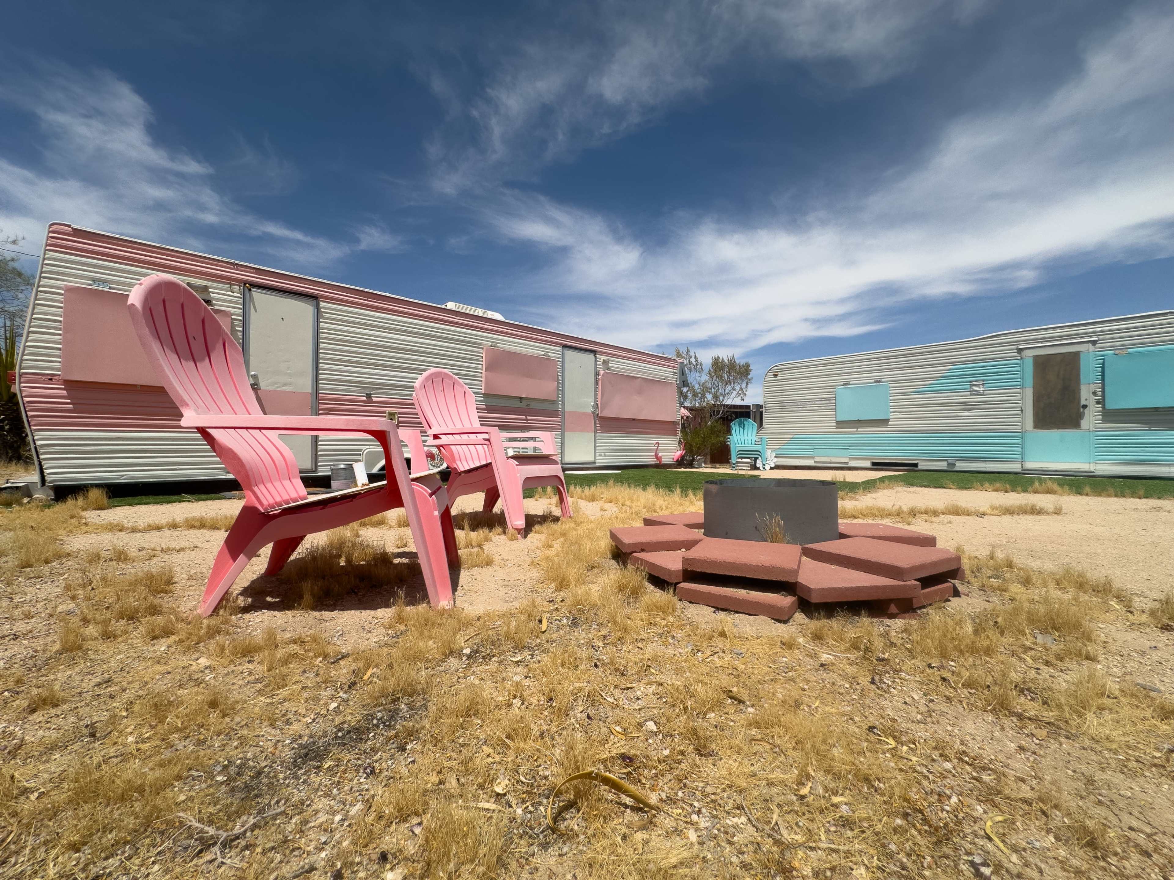 The scene features two retro travel trailers surrounded by a circle of pink Adirondack chairs and a stone fire pit on a dry, grassy area.