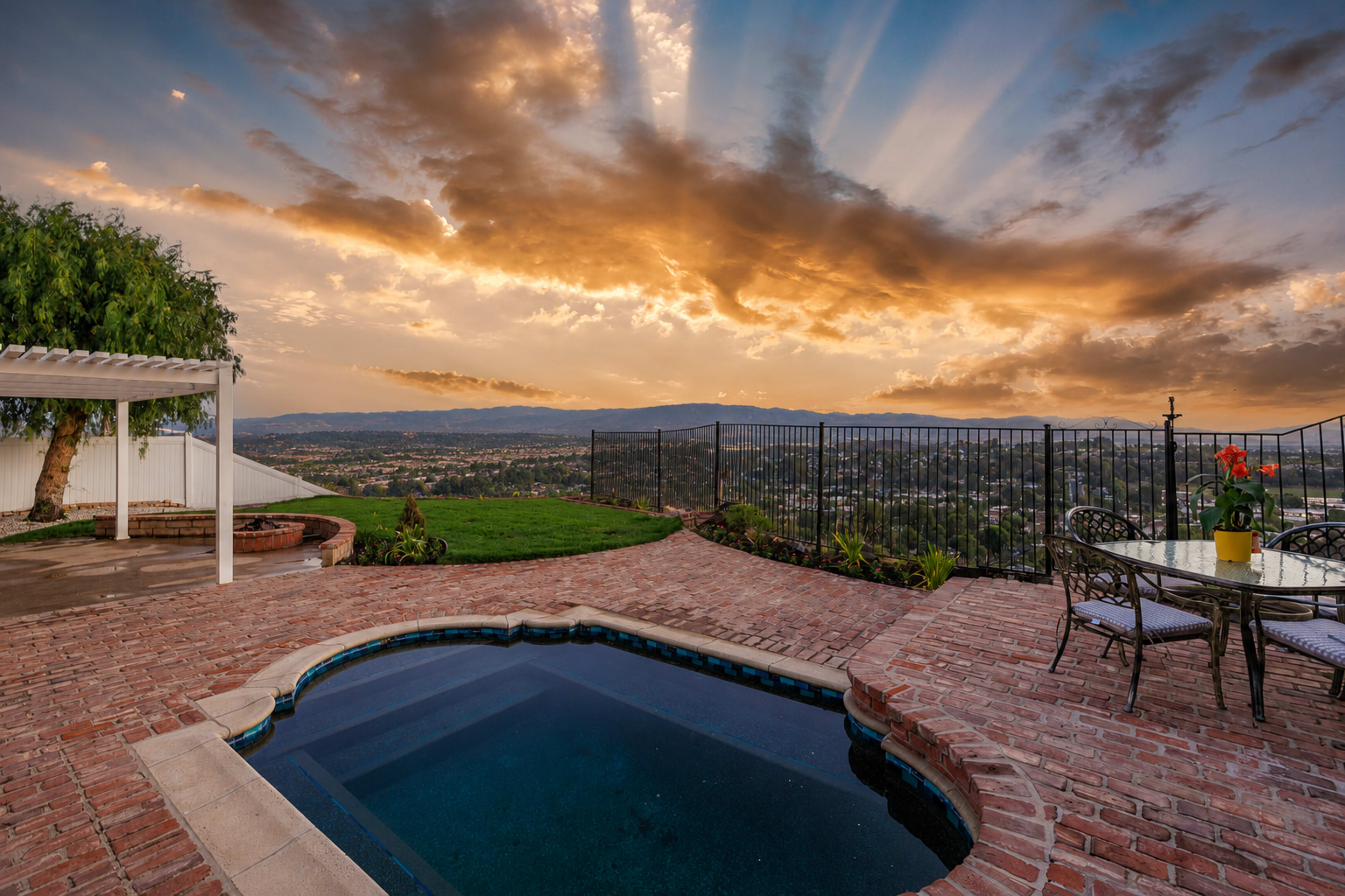 The image shows a backyard scene featuring a hot tub, brick patio, outdoor dining set, and a view of a sunset with dramatic clouds over a valley.