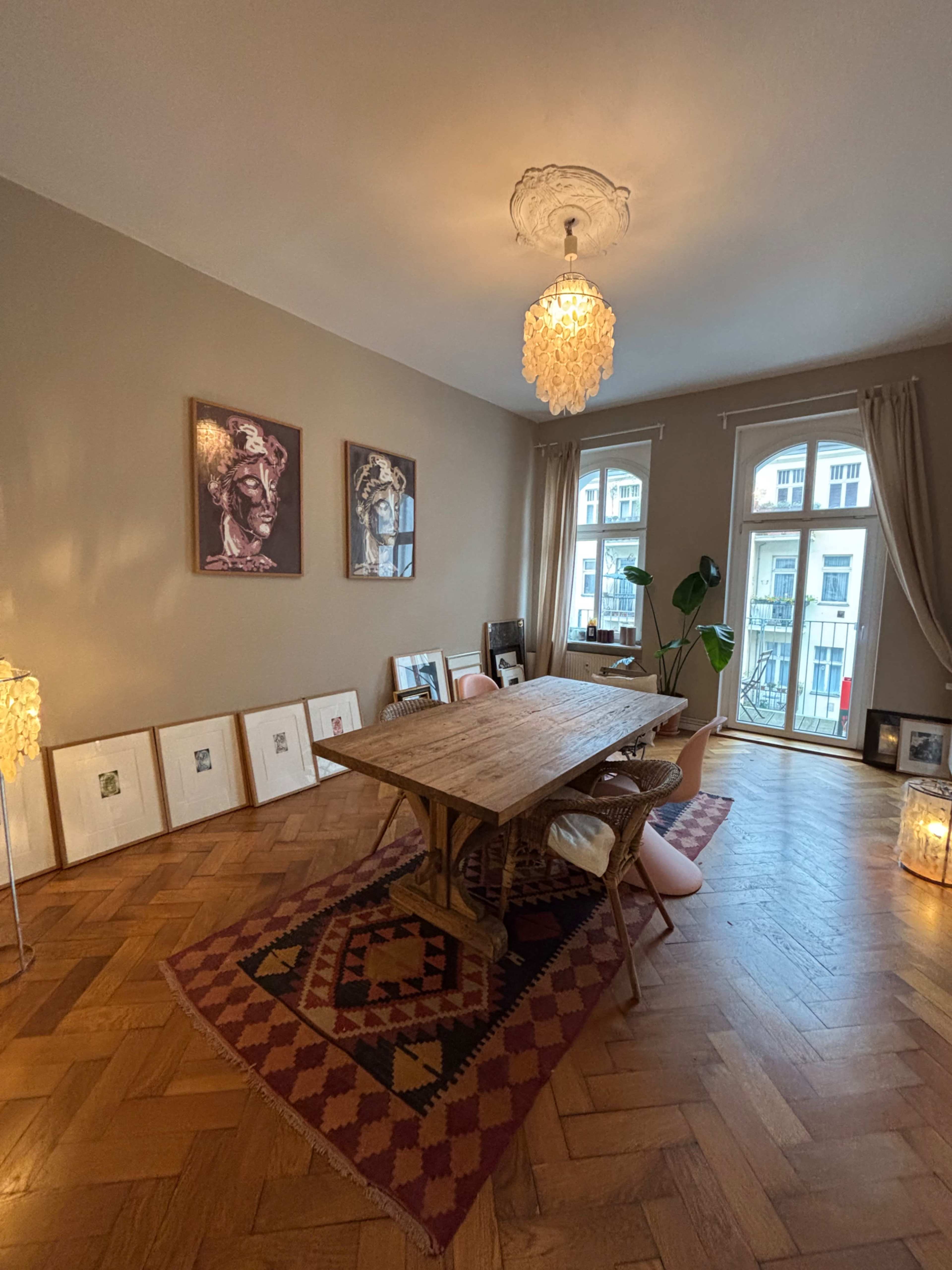 The image shows a well-lit room featuring a wooden dining table surrounded by chairs, framed artwork on the walls, and a patterned rug on a hardwood floor.