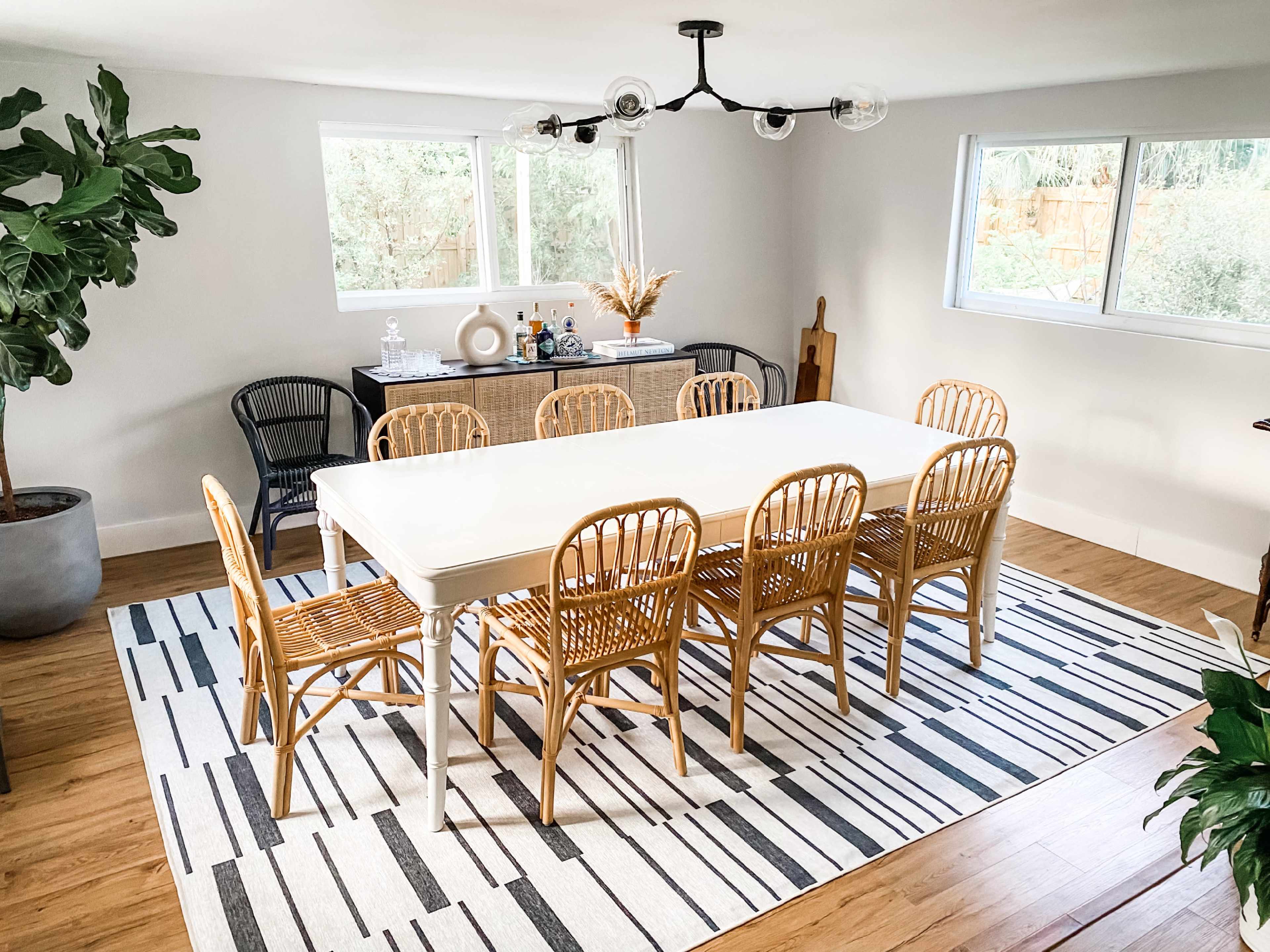 A spacious dining area features a large white table surrounded by six rattan chairs on a striped rug, with a sideboard and large windows in the background.