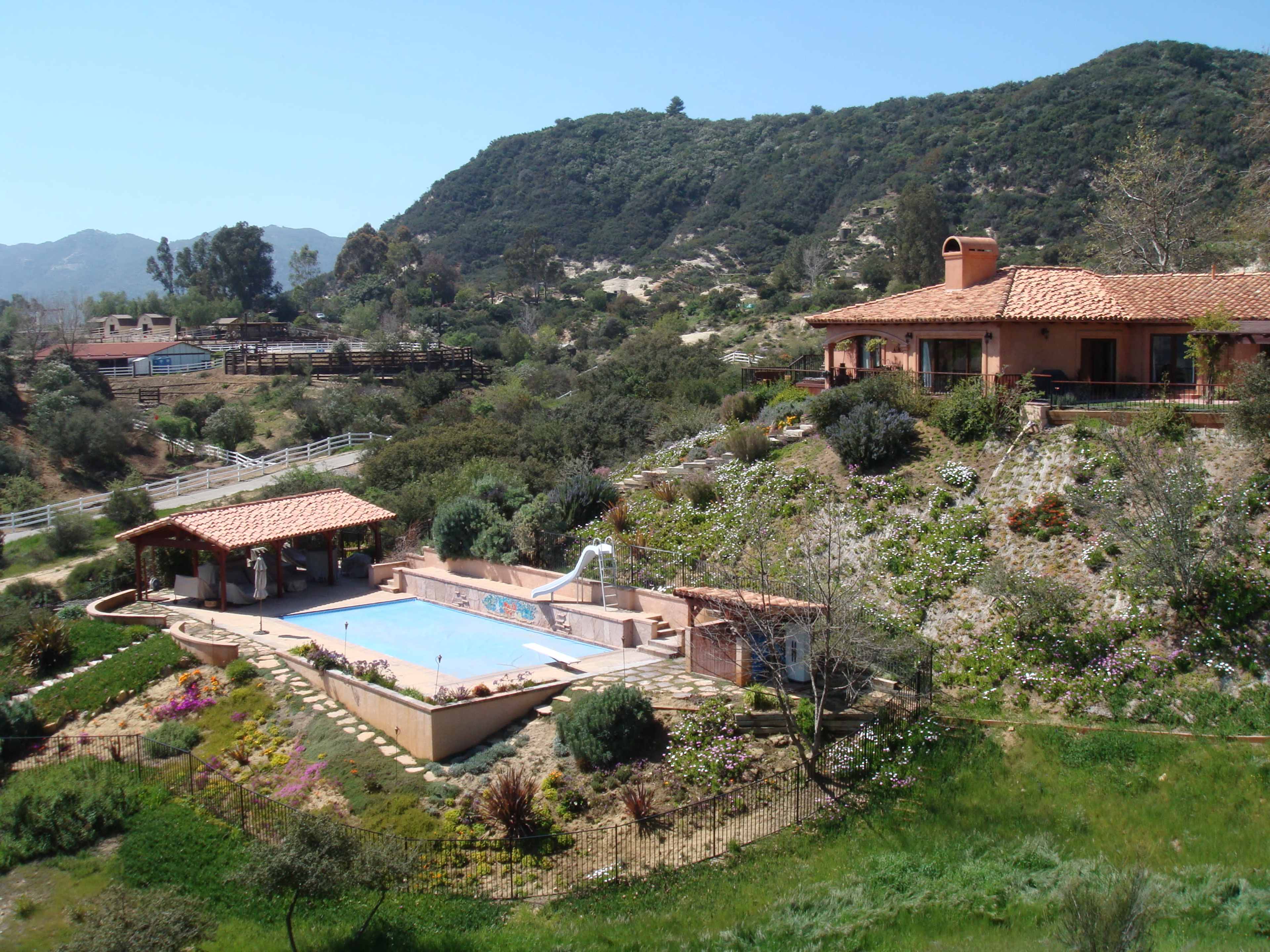The image shows a hillside property featuring a swimming pool with a slide, surrounded by landscaped gardens and distant mountains.