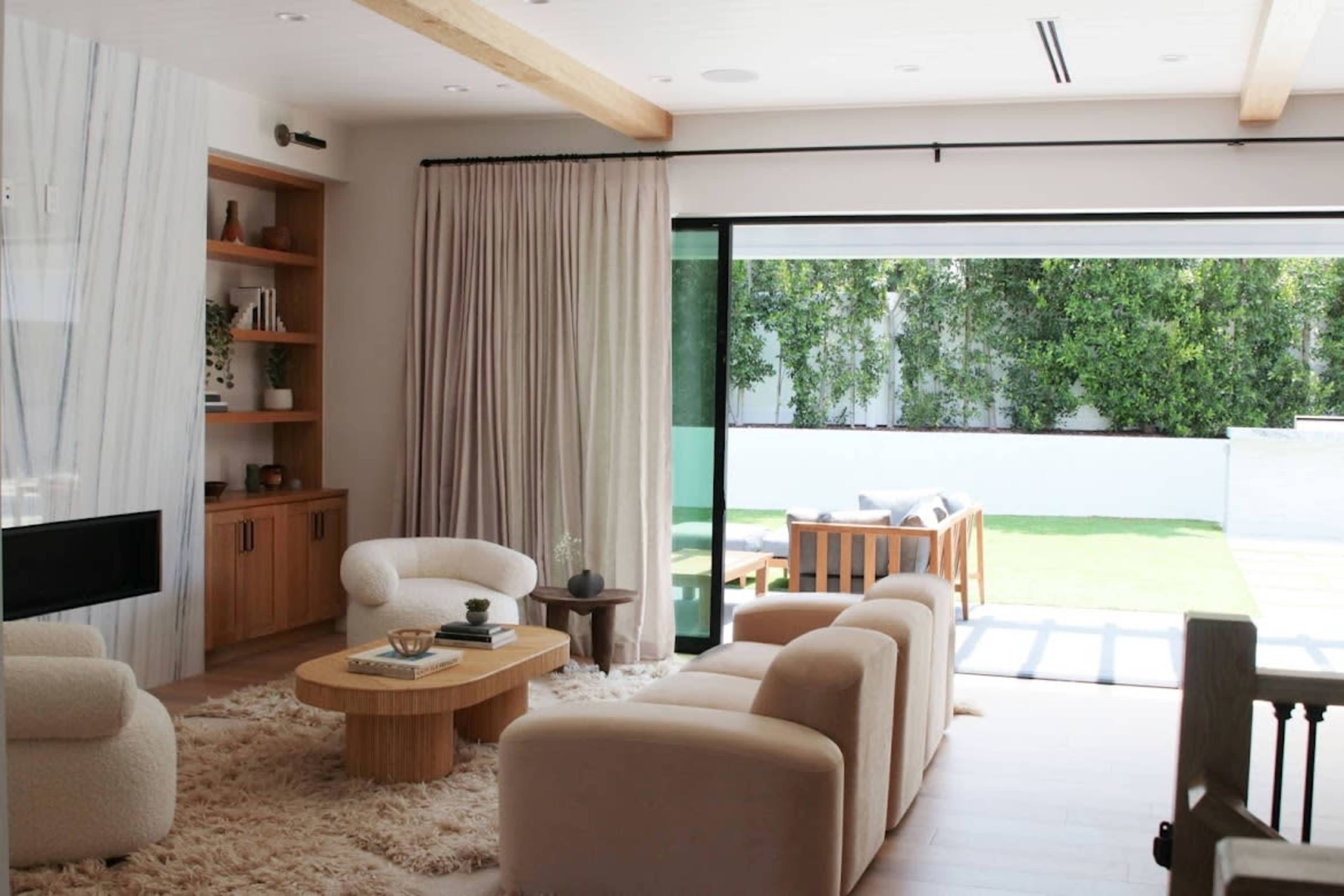 A modern living room features soft furnishings, a round wooden coffee table, and large glass doors opening to a landscaped outdoor area.