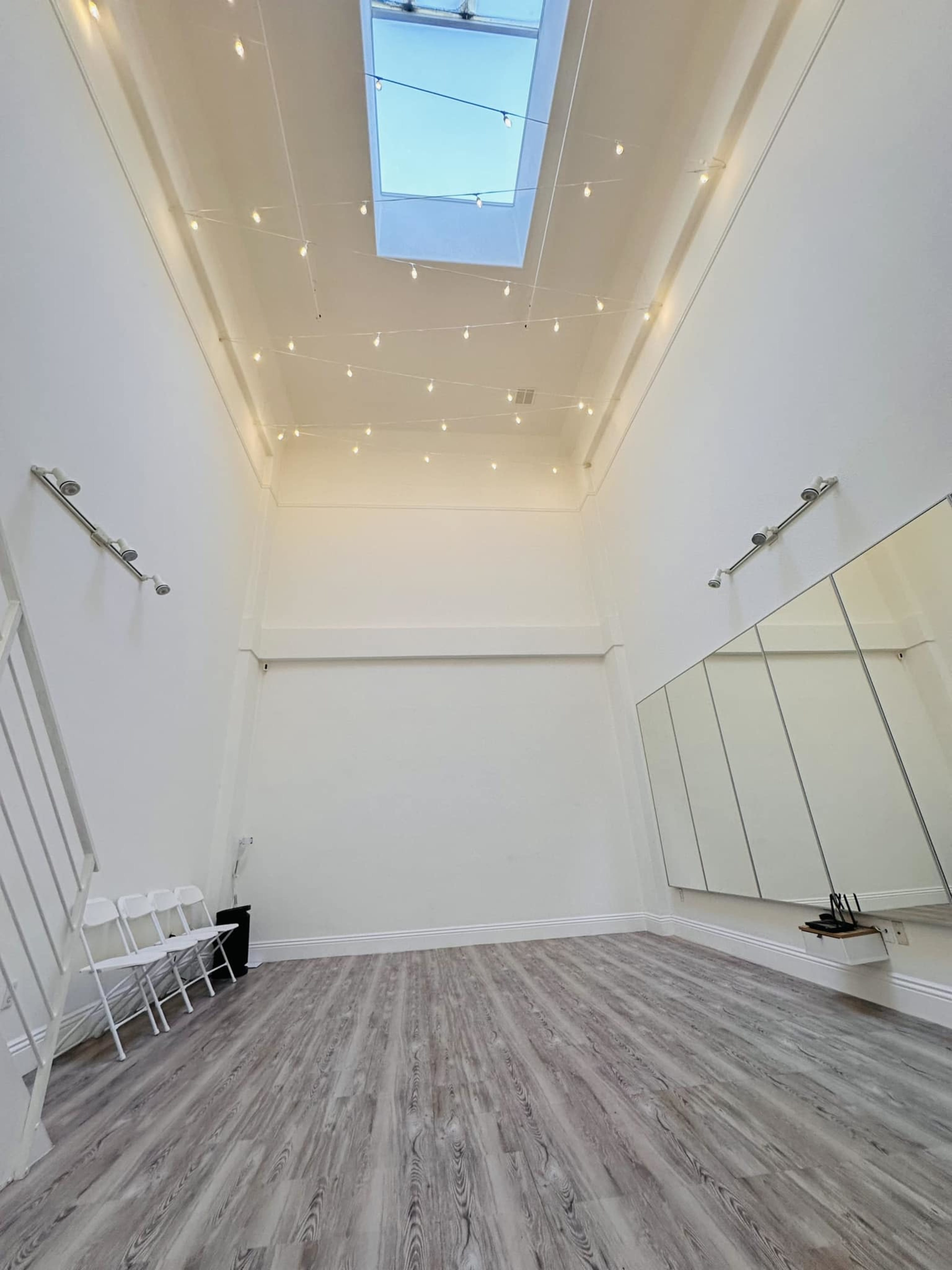 A spacious room with high ceilings, featuring a skylight, string lights, and mirrored walls, along with a row of white chairs in the corner.