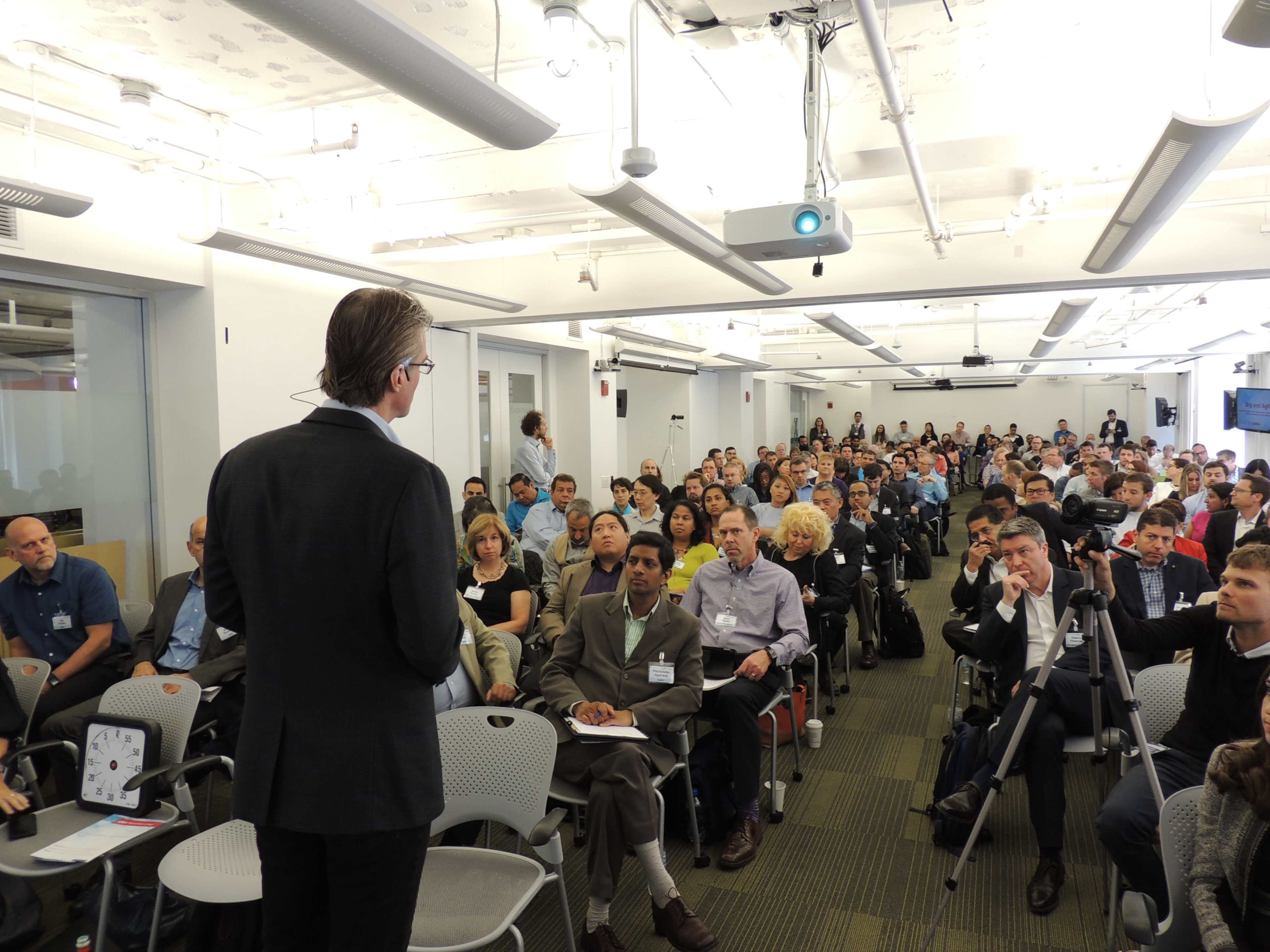 A speaker stands at the front of a large seminar room filled with an audience seated in rows, attentively facing the stage.