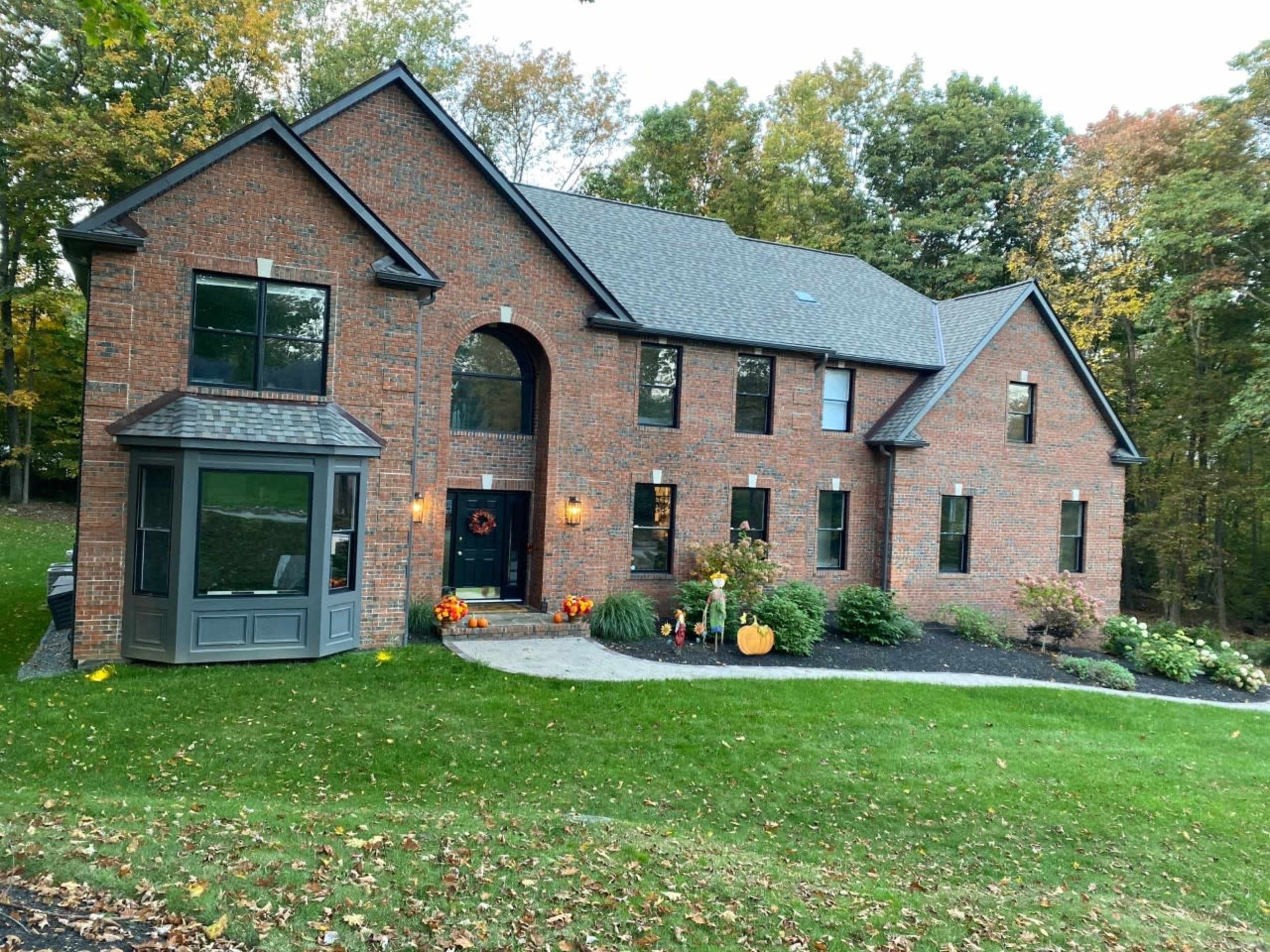 A two-story brick house with large windows is surrounded by a well-kept lawn and autumn decorations, including pumpkins and orange flowers.