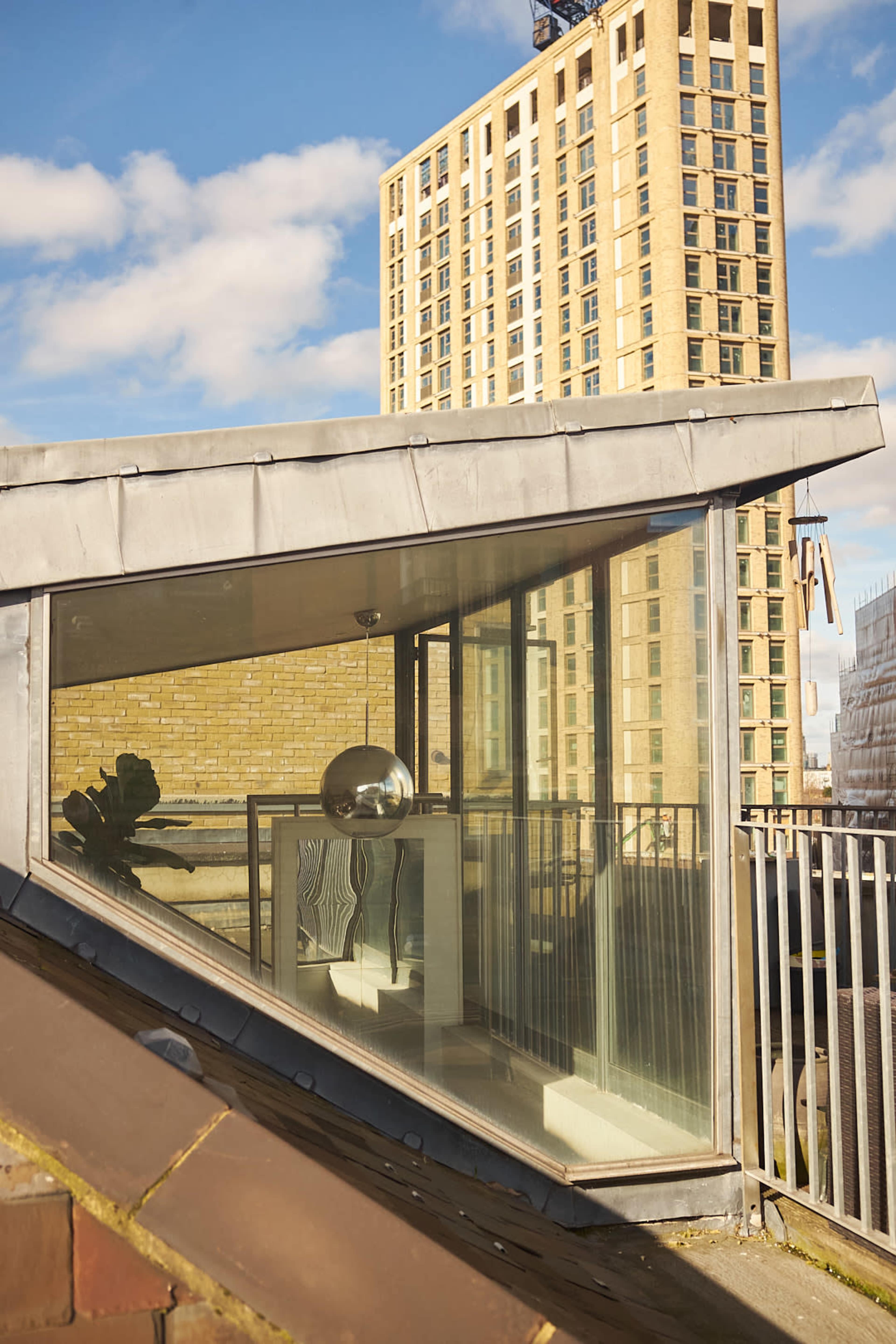 A modern glass-enclosed balcony is positioned on a sloped rooftop, overlooking a tall residential building against a blue sky with scattered clouds.