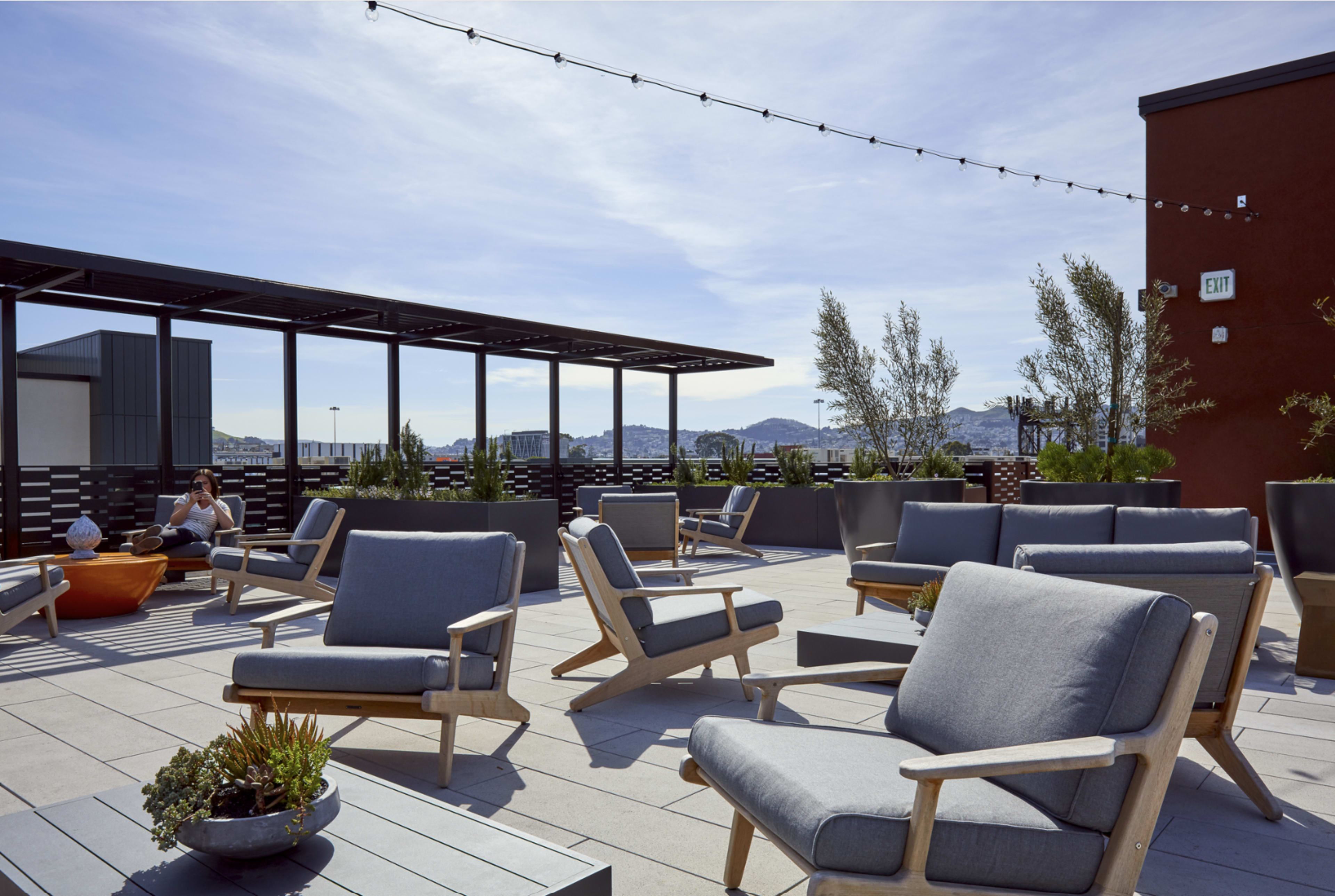 The image shows a modern rooftop terrace with several gray lounge chairs, potted plants, and a scenic view of distant hills under a partly cloudy sky.