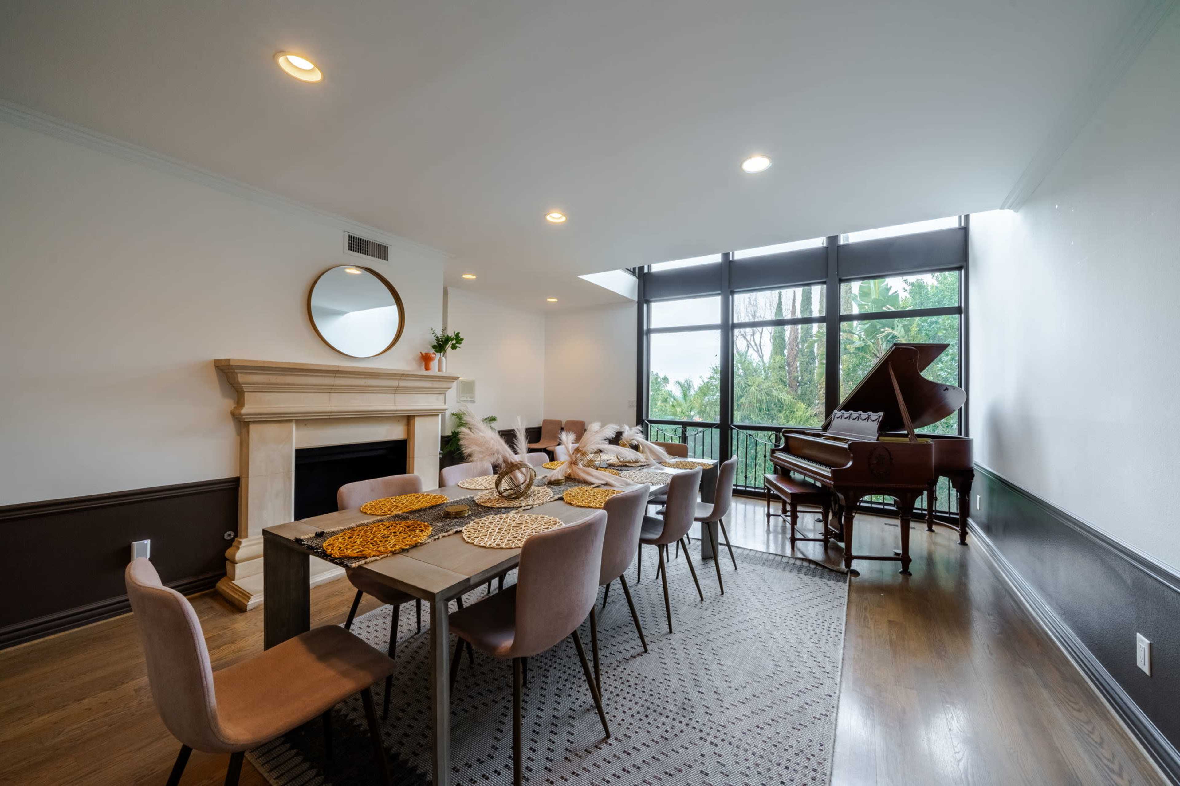 A dining area features a wooden table set with placemats and chairs, alongside a fireplace and a grand piano near large windows.