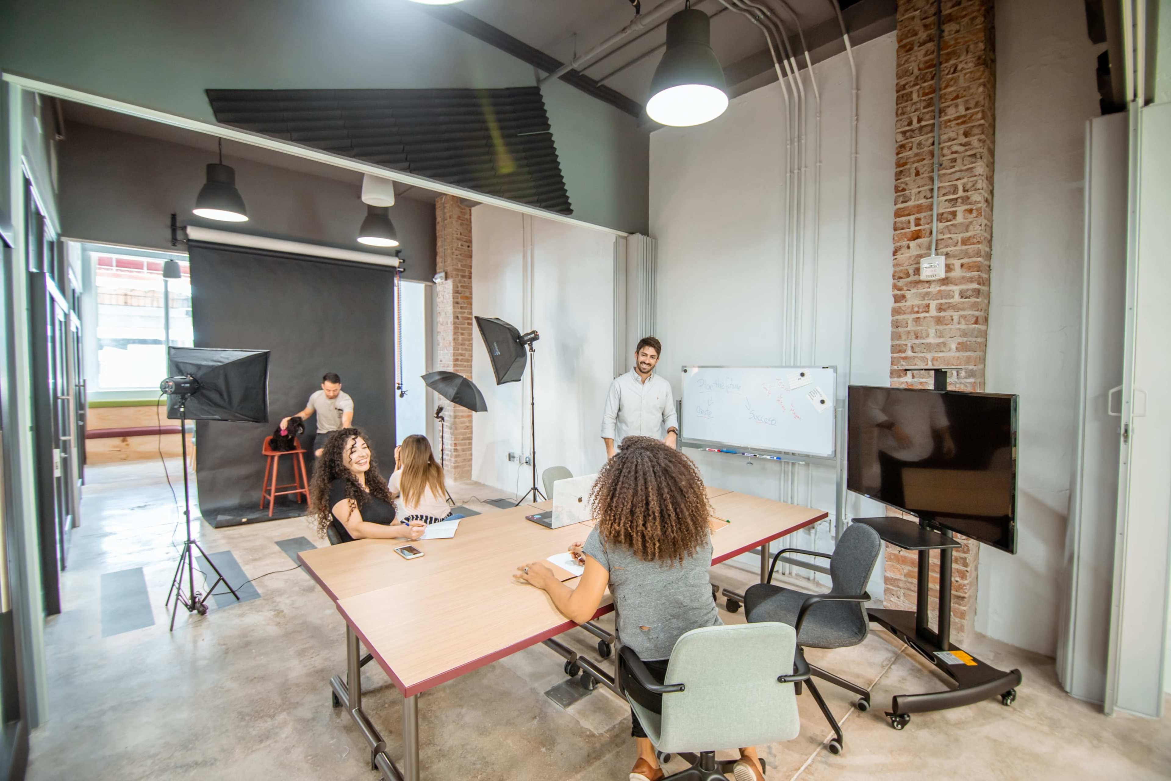 A group of people collaborates around a table in a modern office space, with a whiteboard and photography equipment in the background.