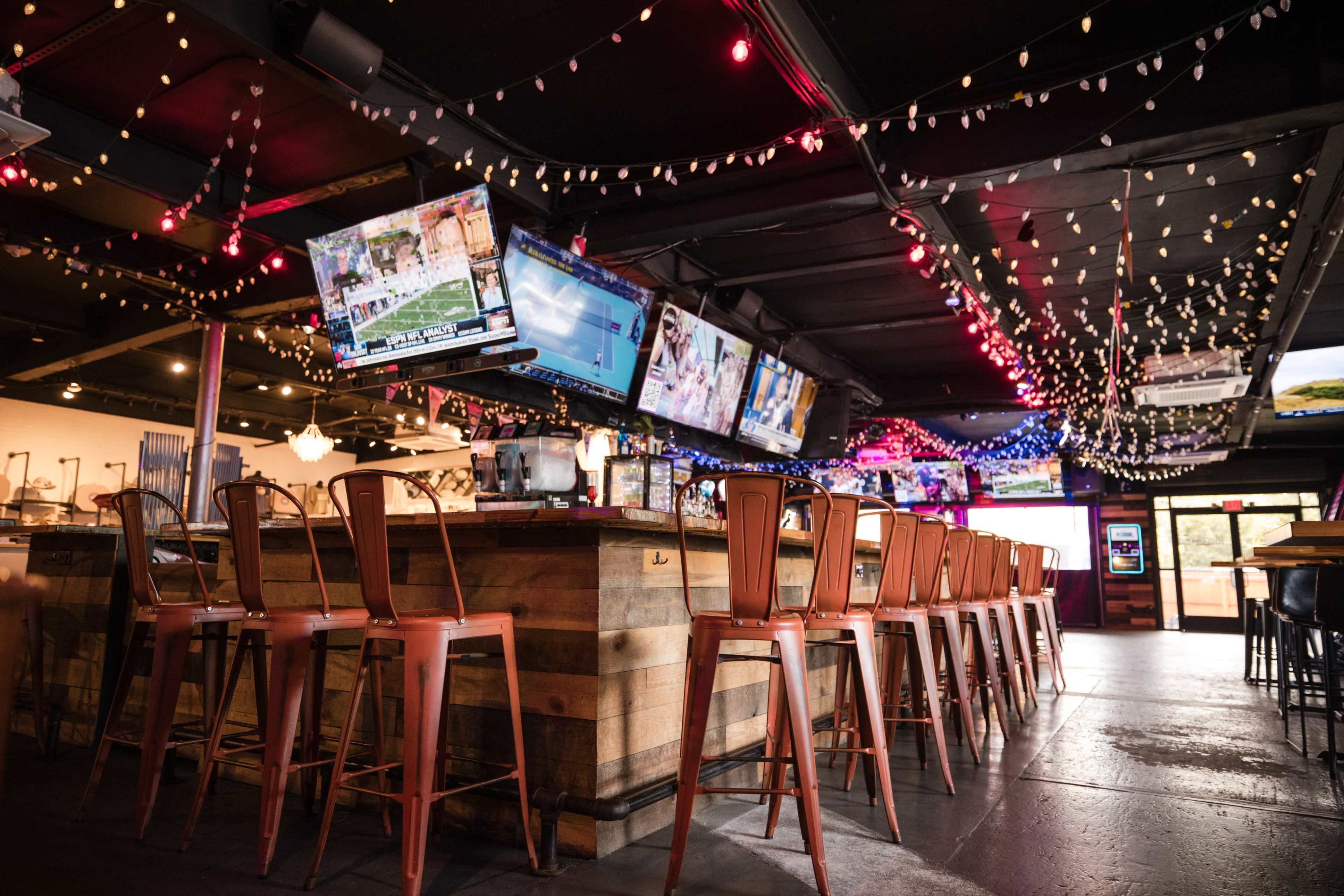 The image shows a bar area with metal stools, multiple television screens displaying sports, and decorative string lights on the ceiling.