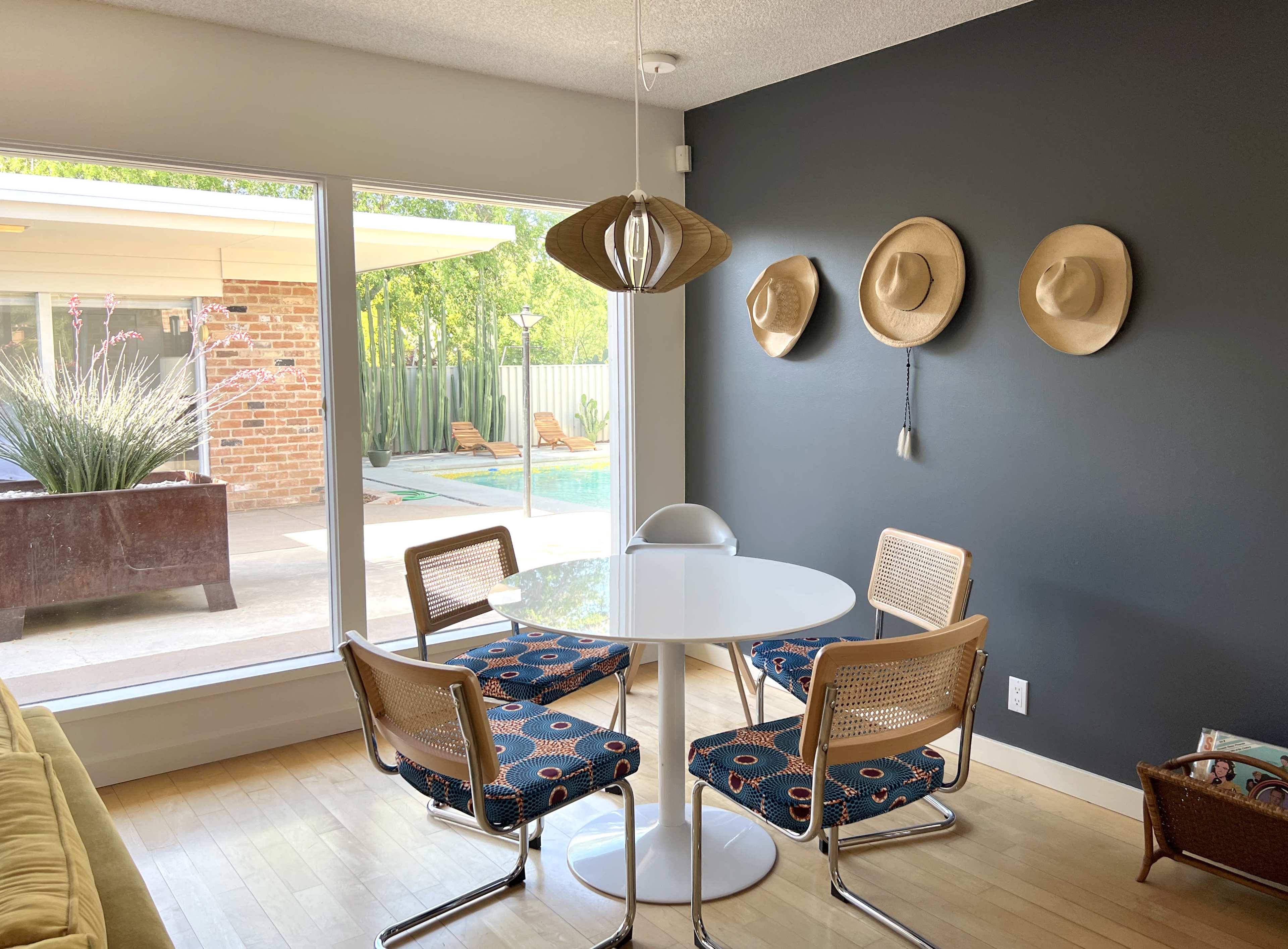 A round white table surrounded by four chairs is set in a room with a large window overlooking a pool and patio area, while three hats are hung on the dark wall.