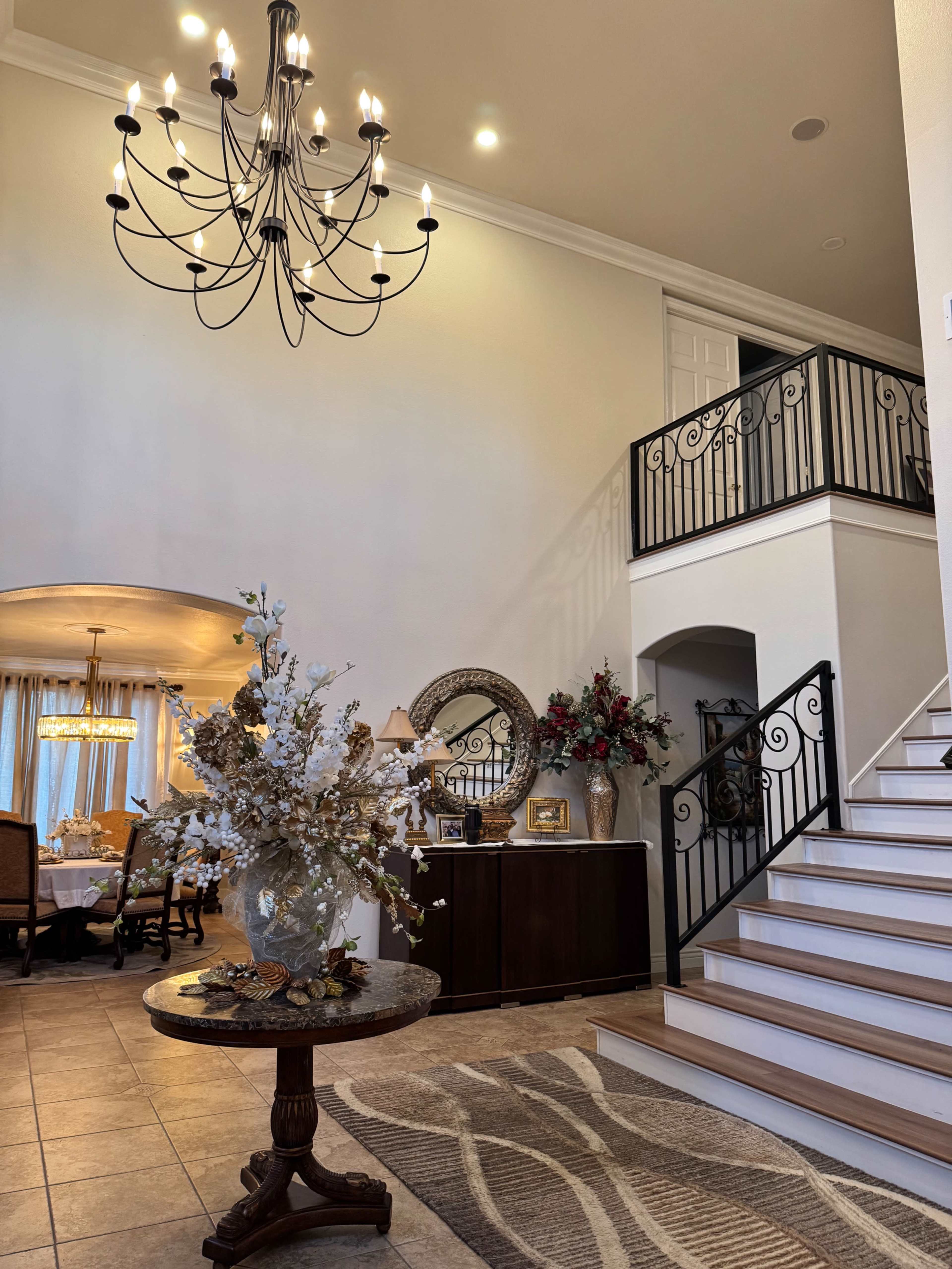 The image shows a spacious entryway featuring a staircase with decorative railings, a chandelier, and a table adorned with floral arrangements.