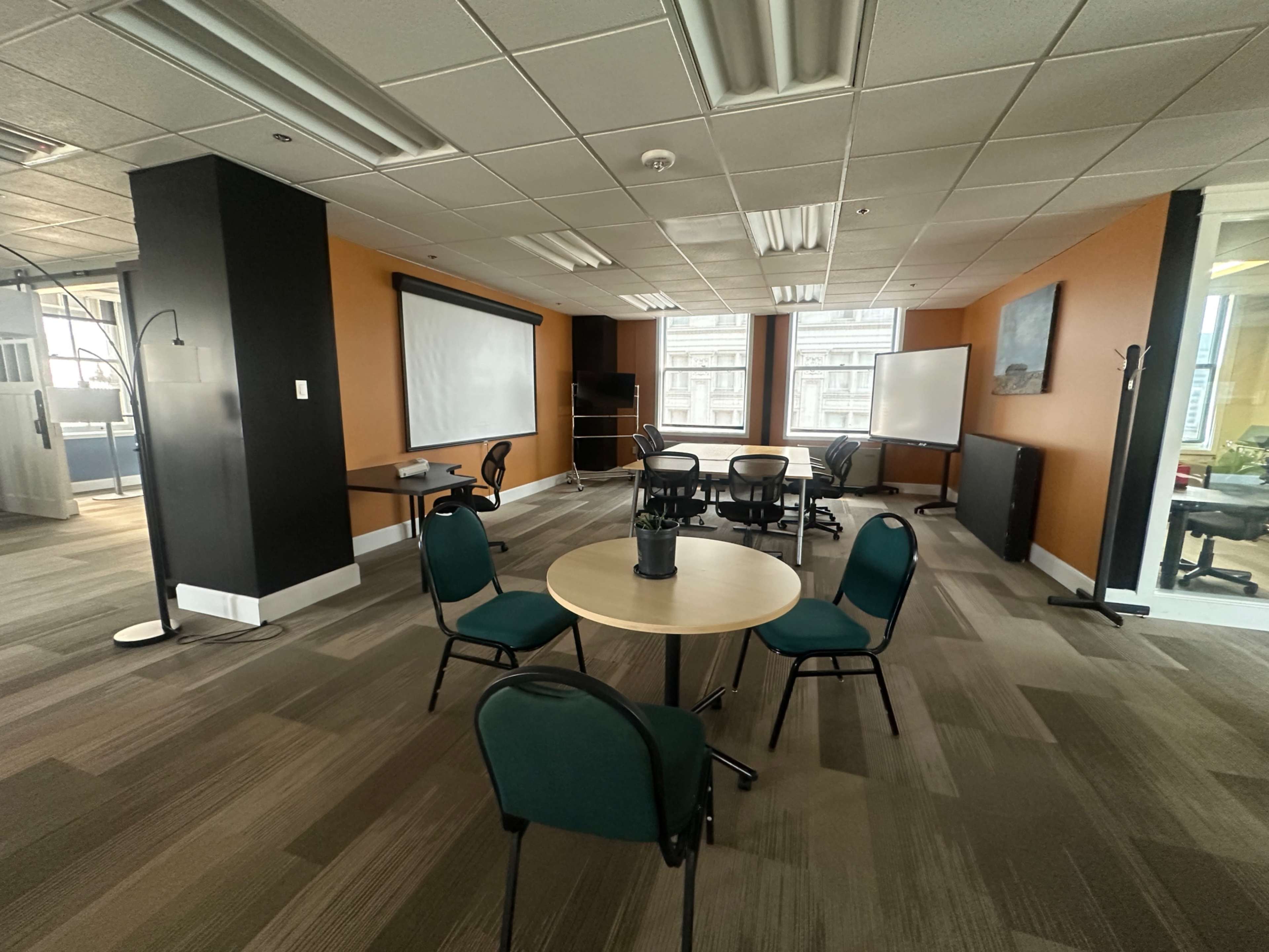The image shows a modern office space with a circular table surrounded by chairs, a projector screen, and multiple windows.