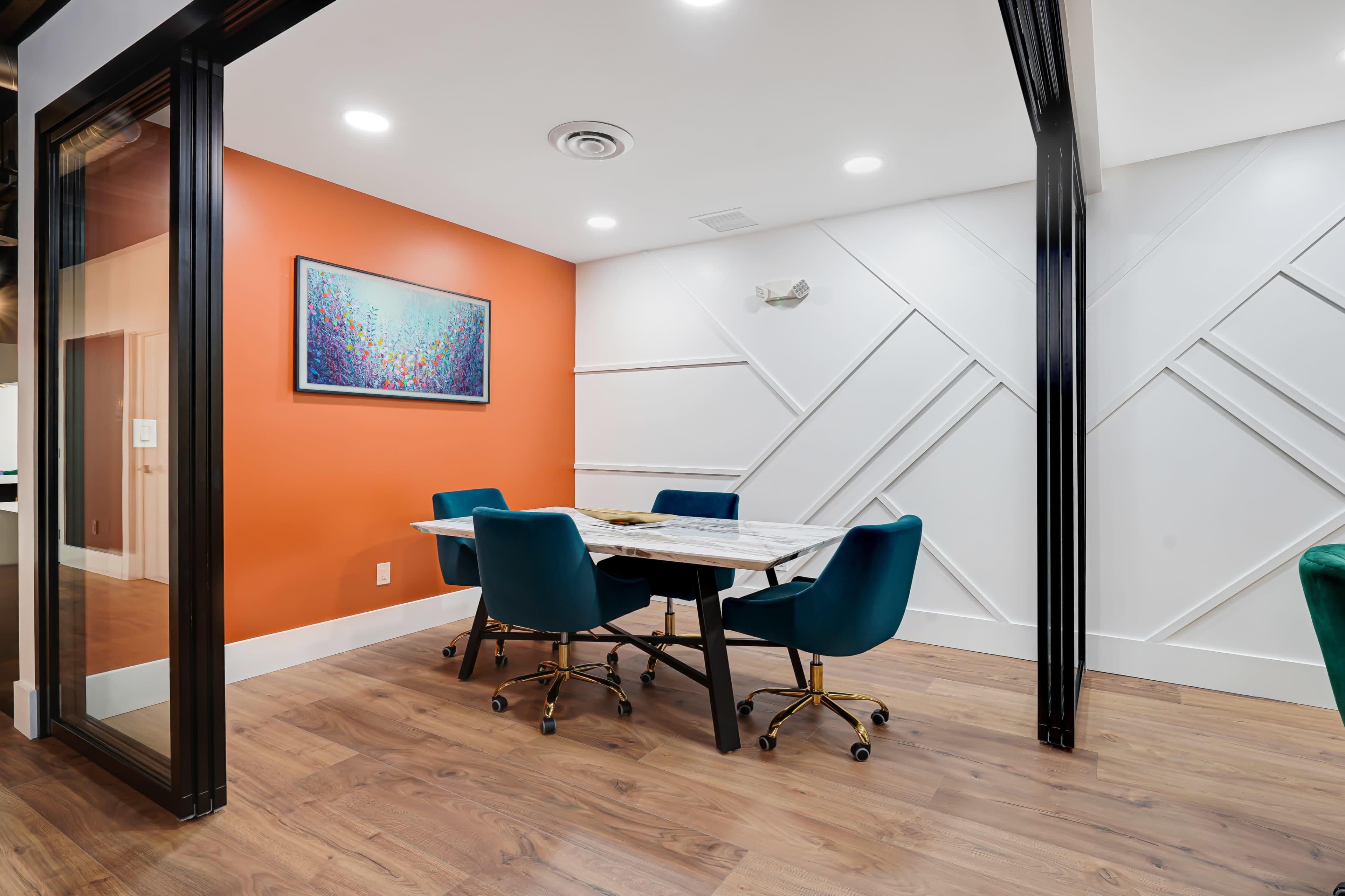 A modern meeting room features a marble table surrounded by teal chairs against an orange accent wall with abstract artwork.