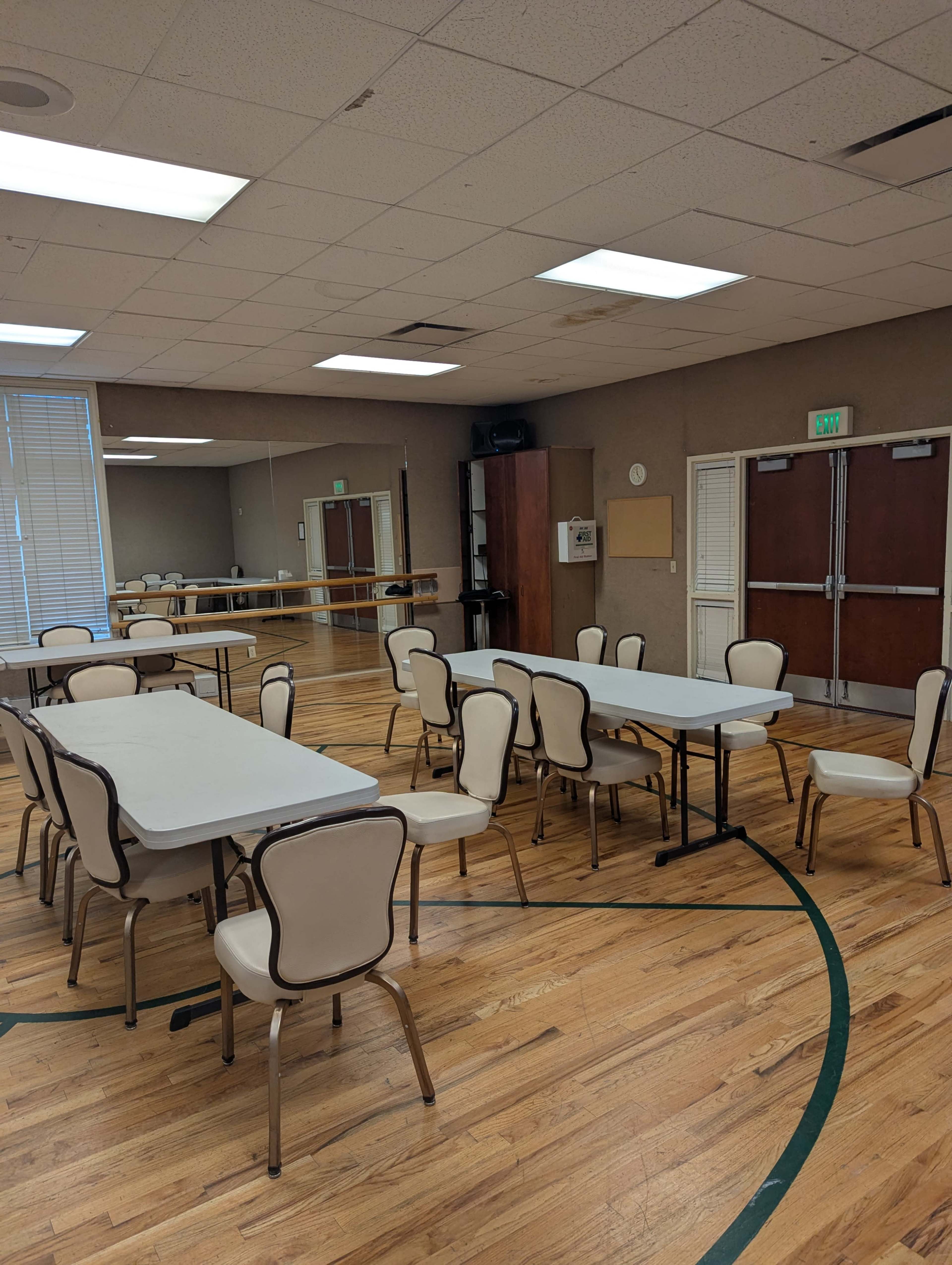 The image shows a multi-purpose room with several tables and chairs arranged around a wooden floor, and mirrors on one wall.