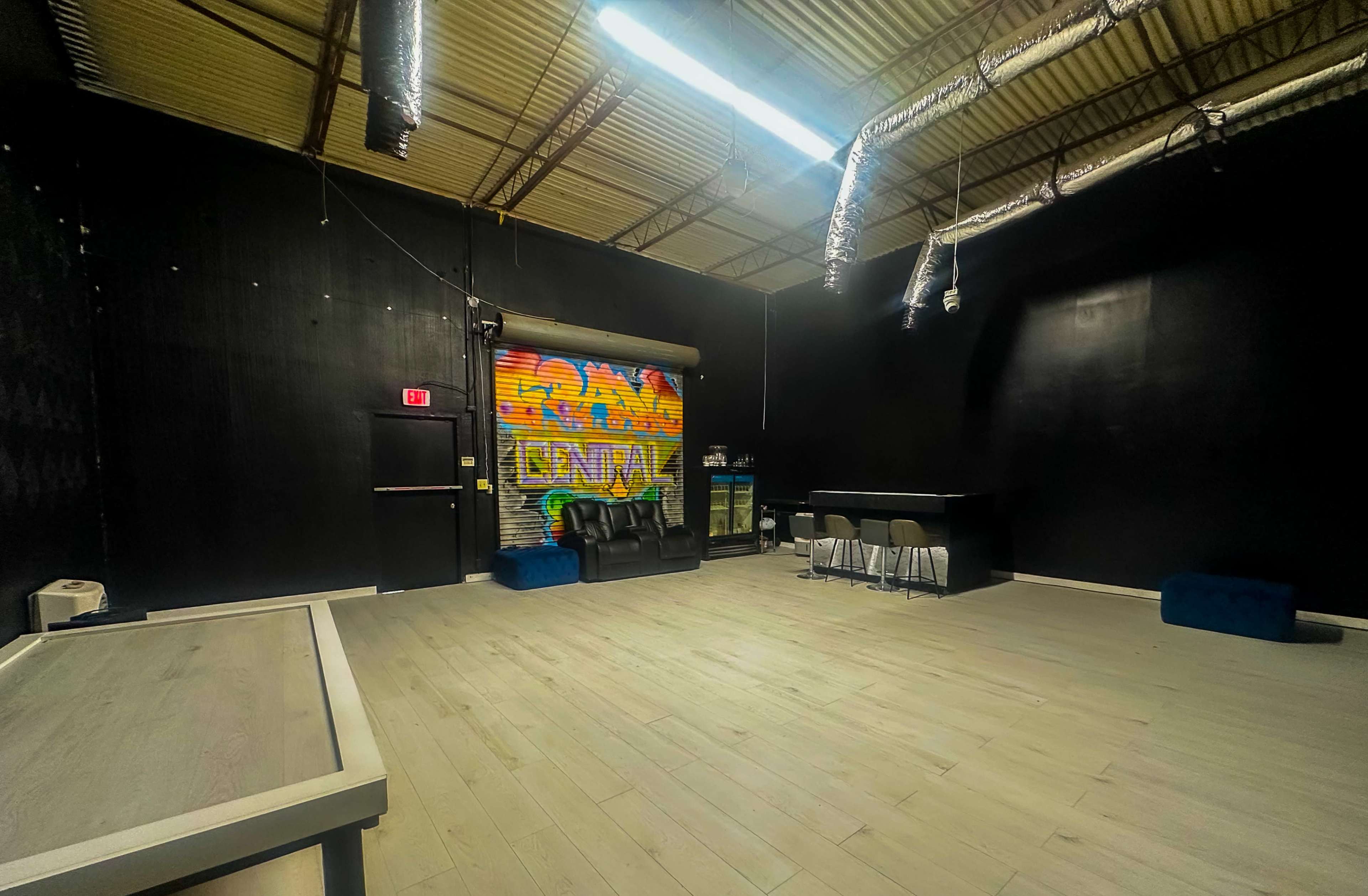 The image shows an empty industrial-style room with black walls, wooden flooring, and a colorful graffiti mural on one wall, along with seating and a small bar area.