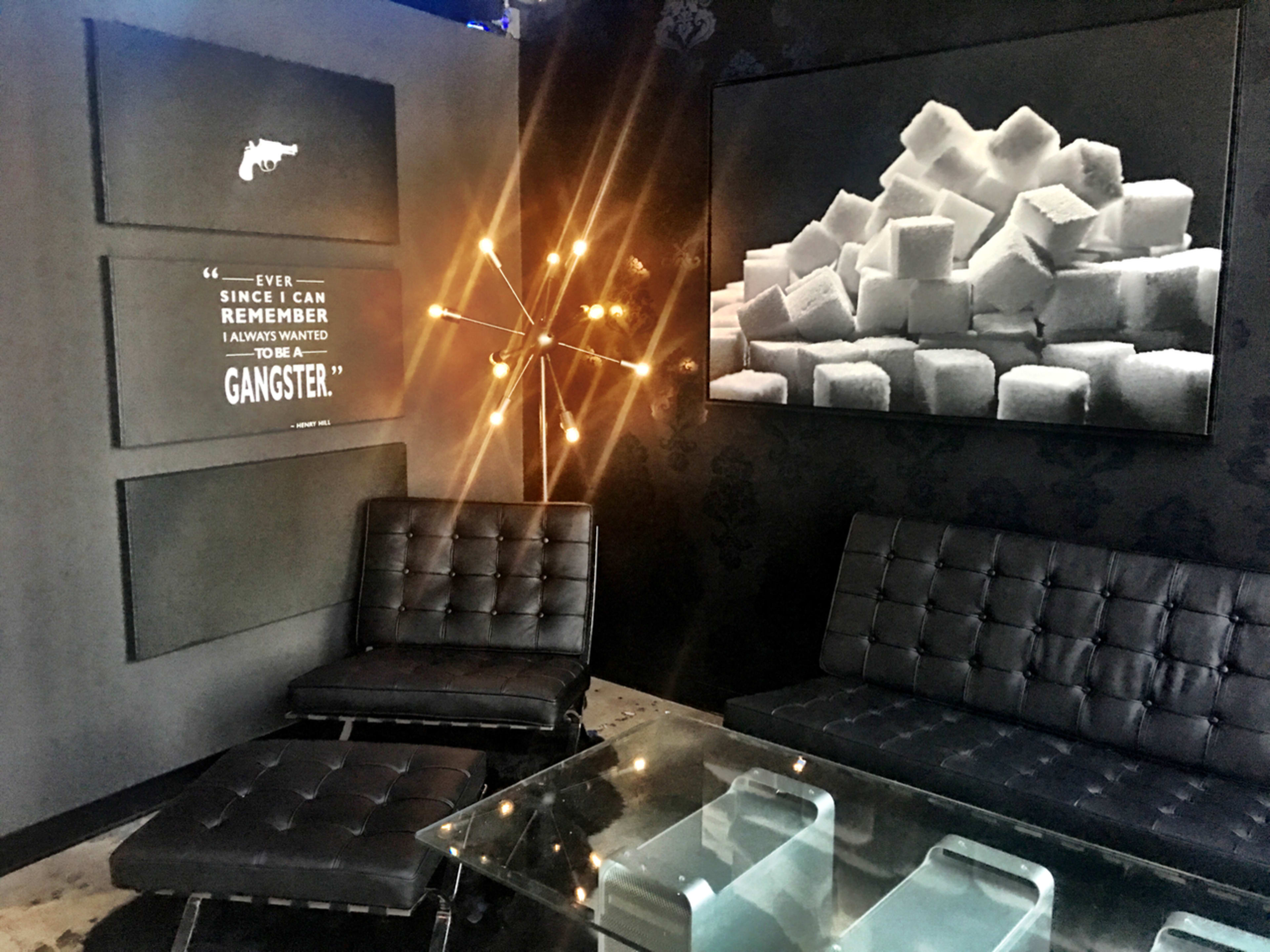 The image shows a modern interior with two black leather chairs, a glass table, and decorative wall art featuring a pile of sugar cubes and a quote about wanting to be a gangster.
