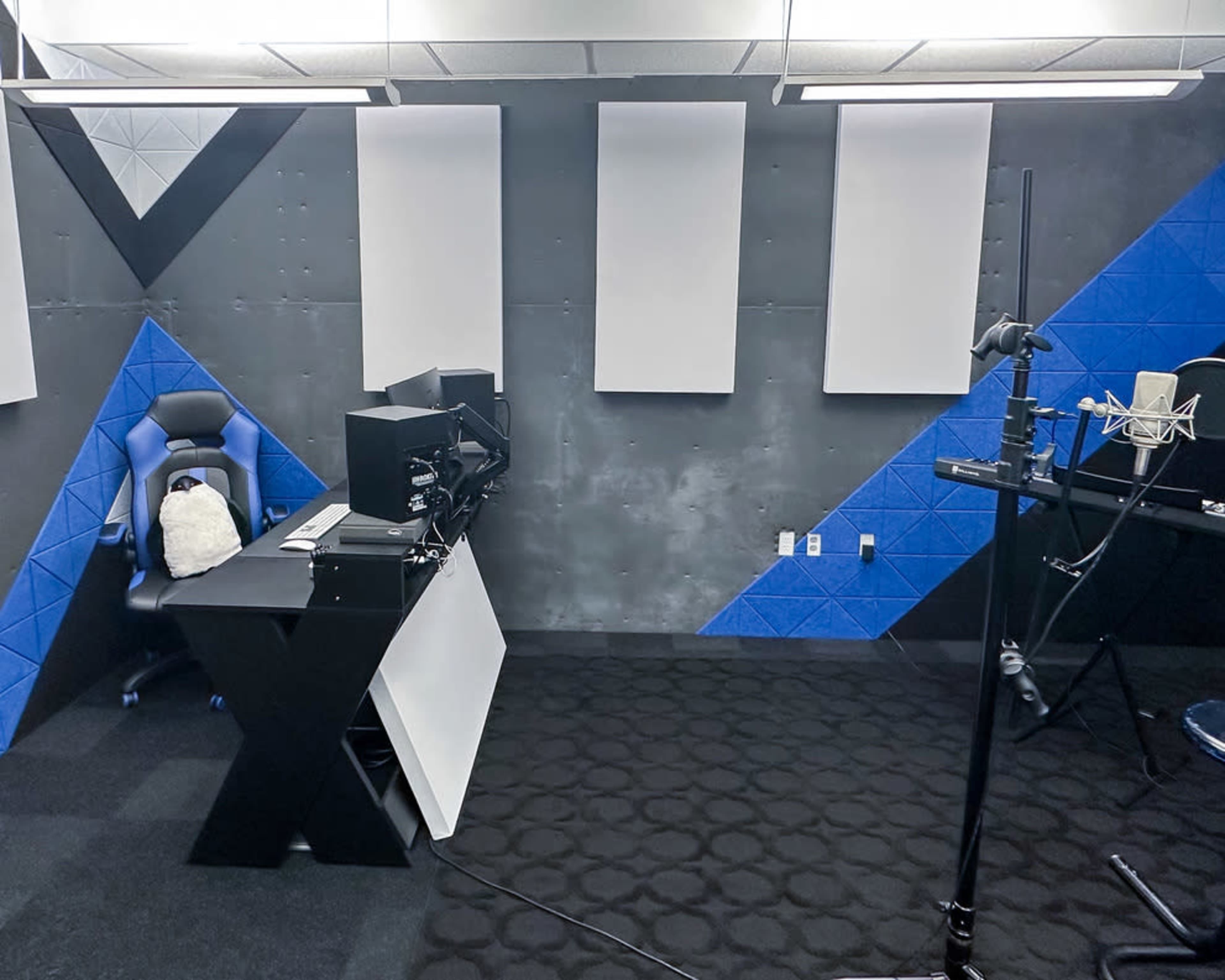 A stylish recording studio with a blue and black color scheme, featuring a microphone on a stand, a gaming chair, and soundproofing panels on the walls.