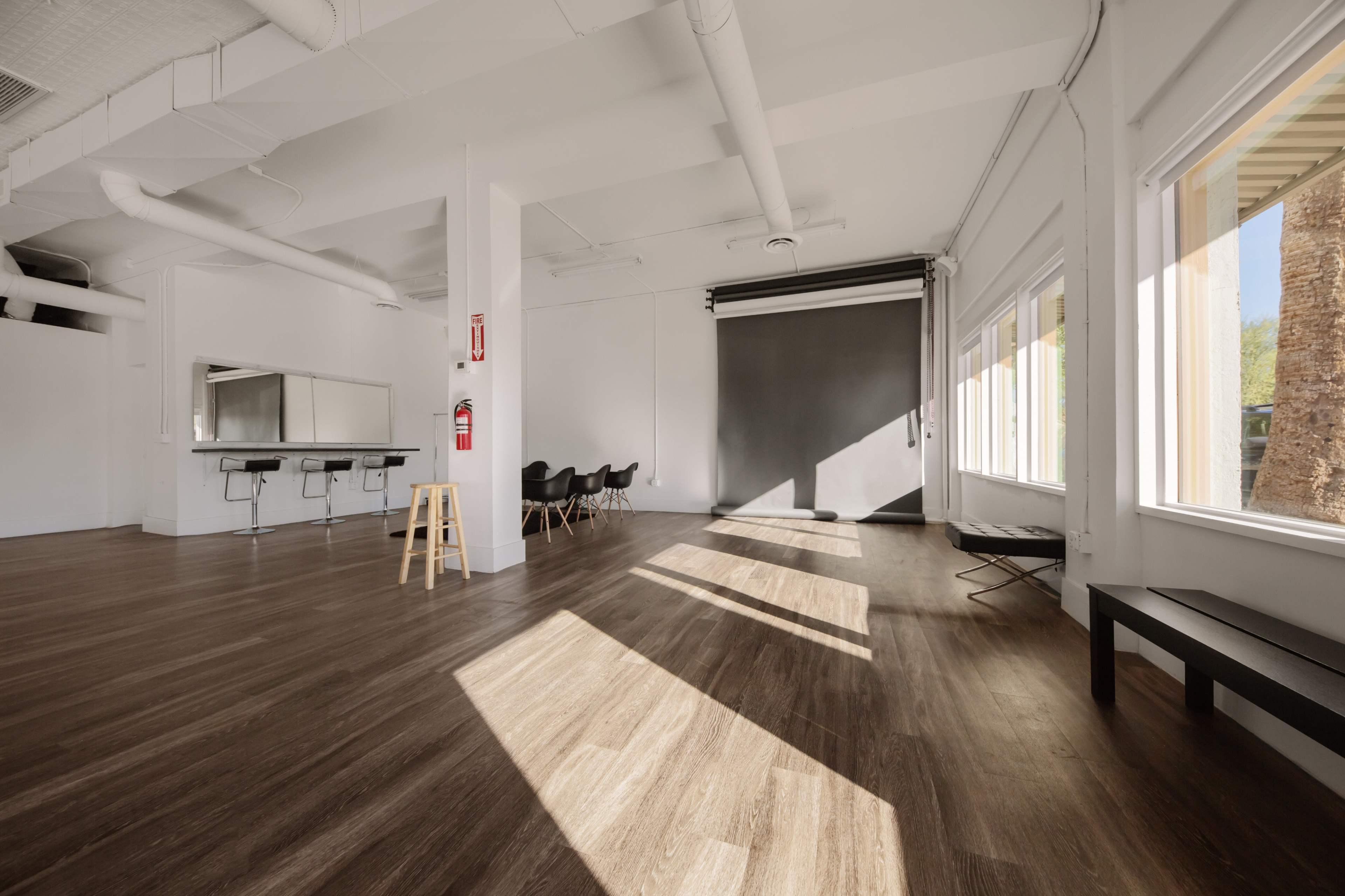 A spacious interior with wooden flooring, featuring large windows, several chairs, and a bar counter against a white wall.