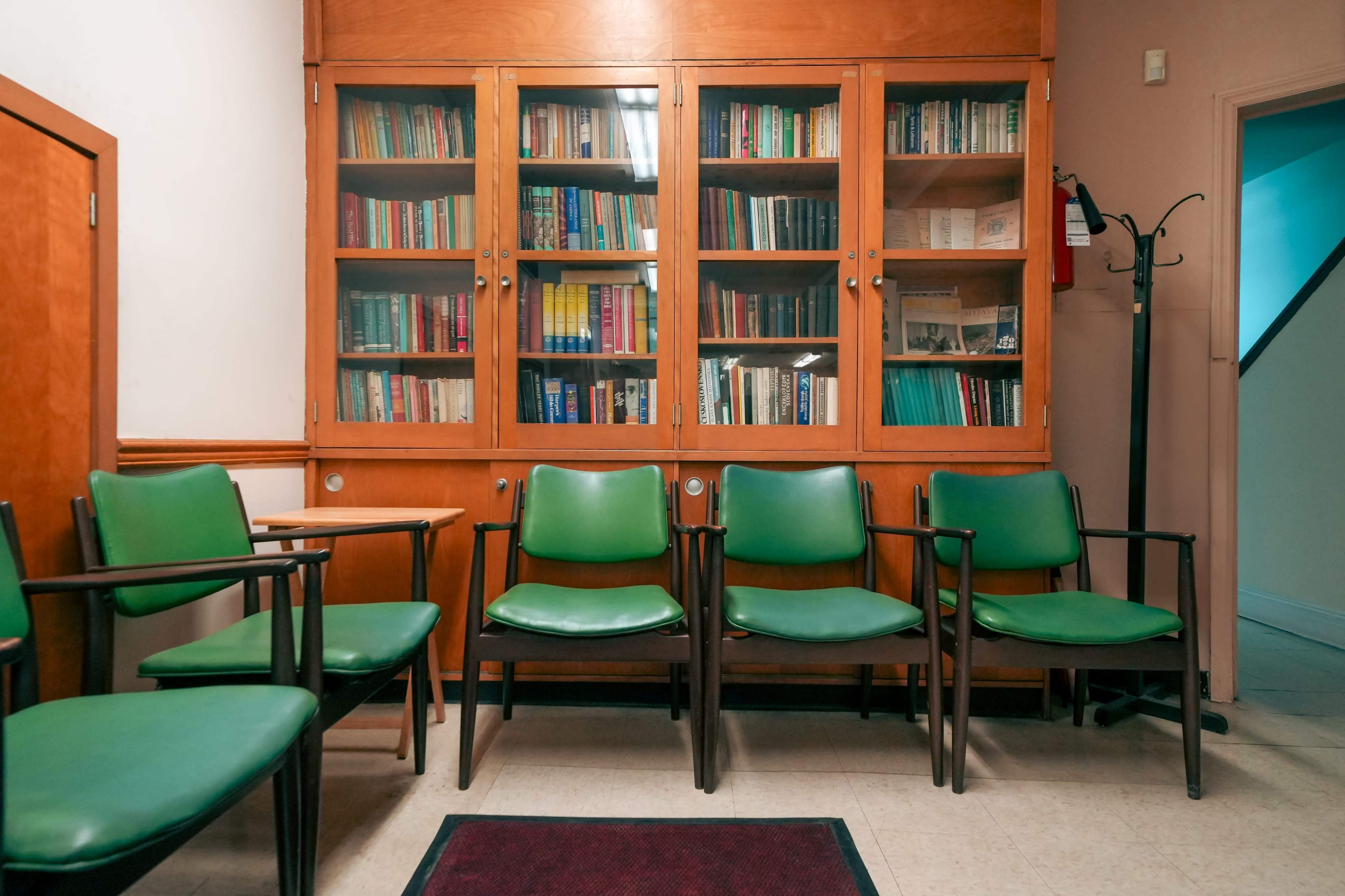 The image shows a waiting area with several green chairs arranged in front of a wooden bookshelf filled with books.