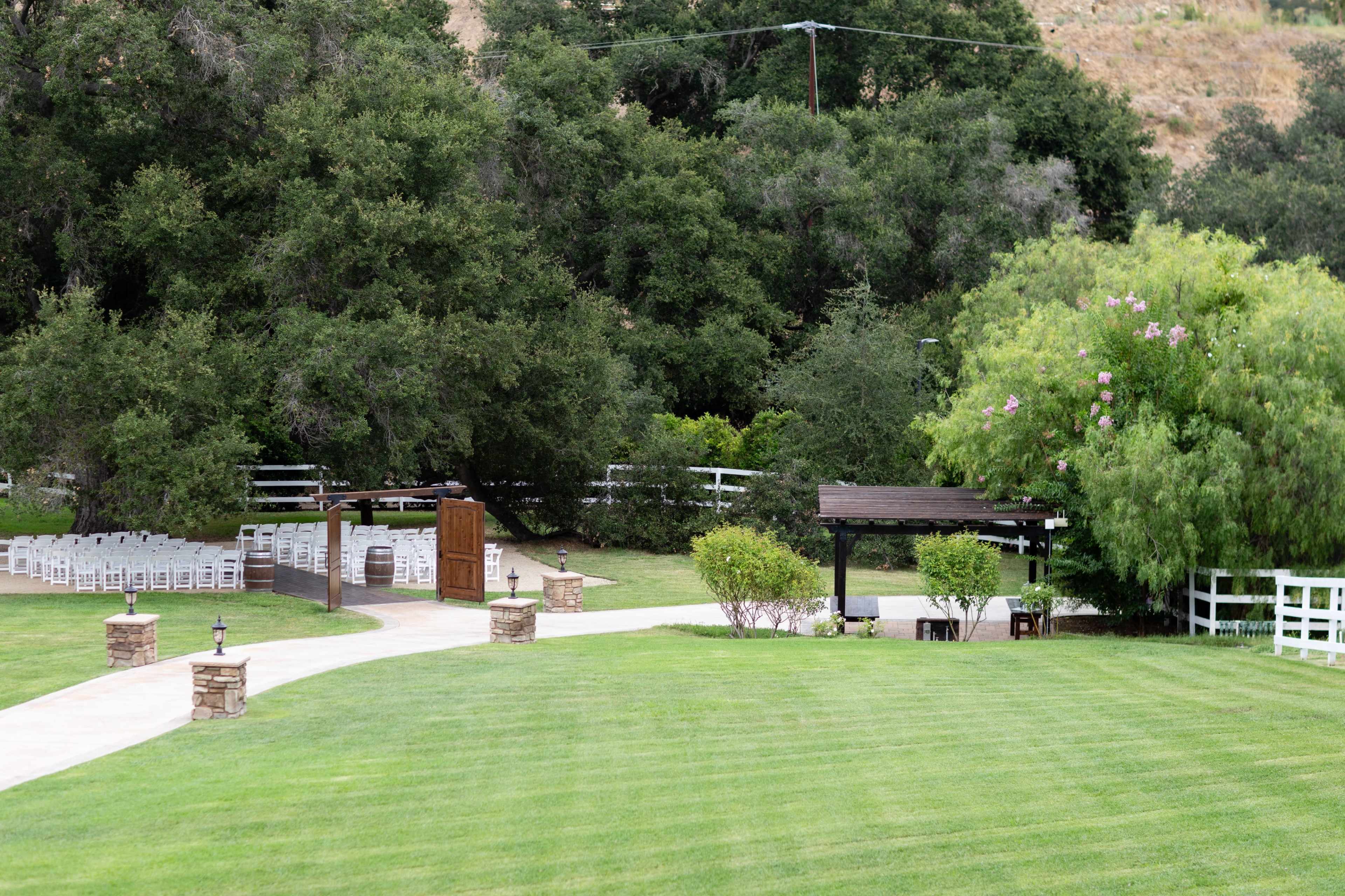 The image shows a landscaped outdoor area with a pathway leading to a ceremony site surrounded by chairs, greenery, and a covered seating area.