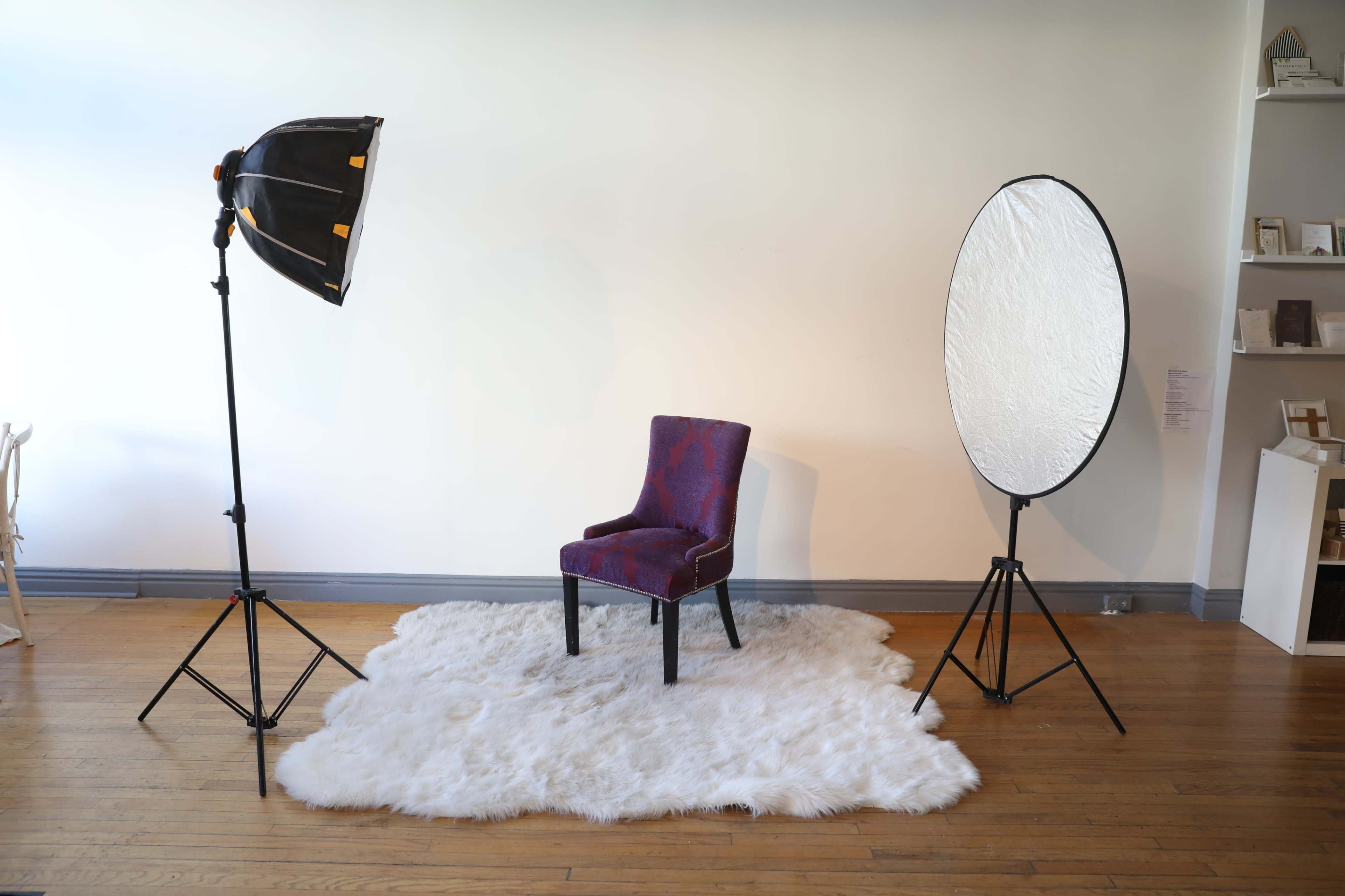 A studio setup featuring a patterned chair placed on a fluffy white rug, illuminated by a softbox light and a large reflector.