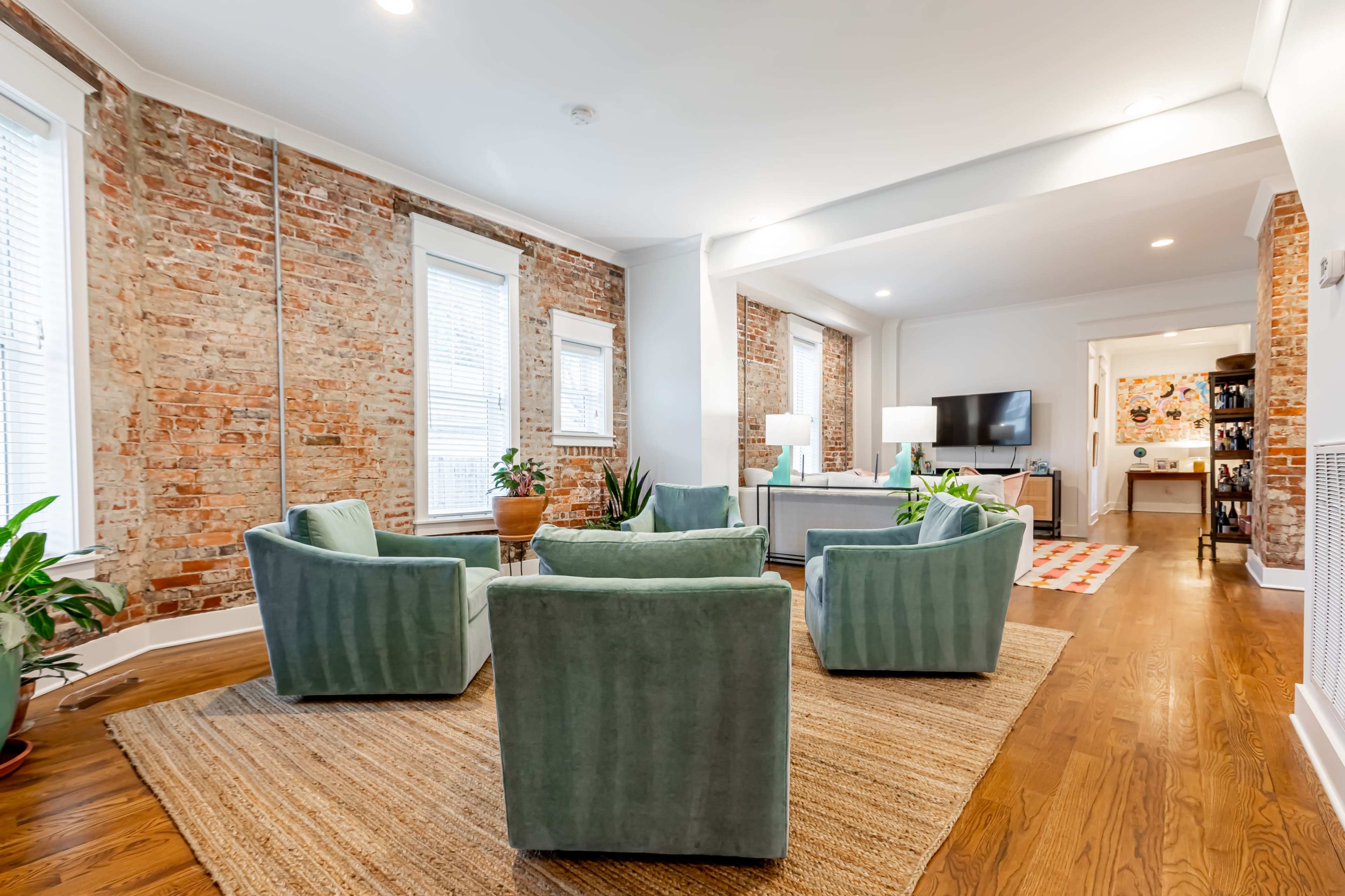 The image shows a living room with exposed brick walls, four teal armchairs arranged in a circular formation, and a large area rug on hardwood flooring.