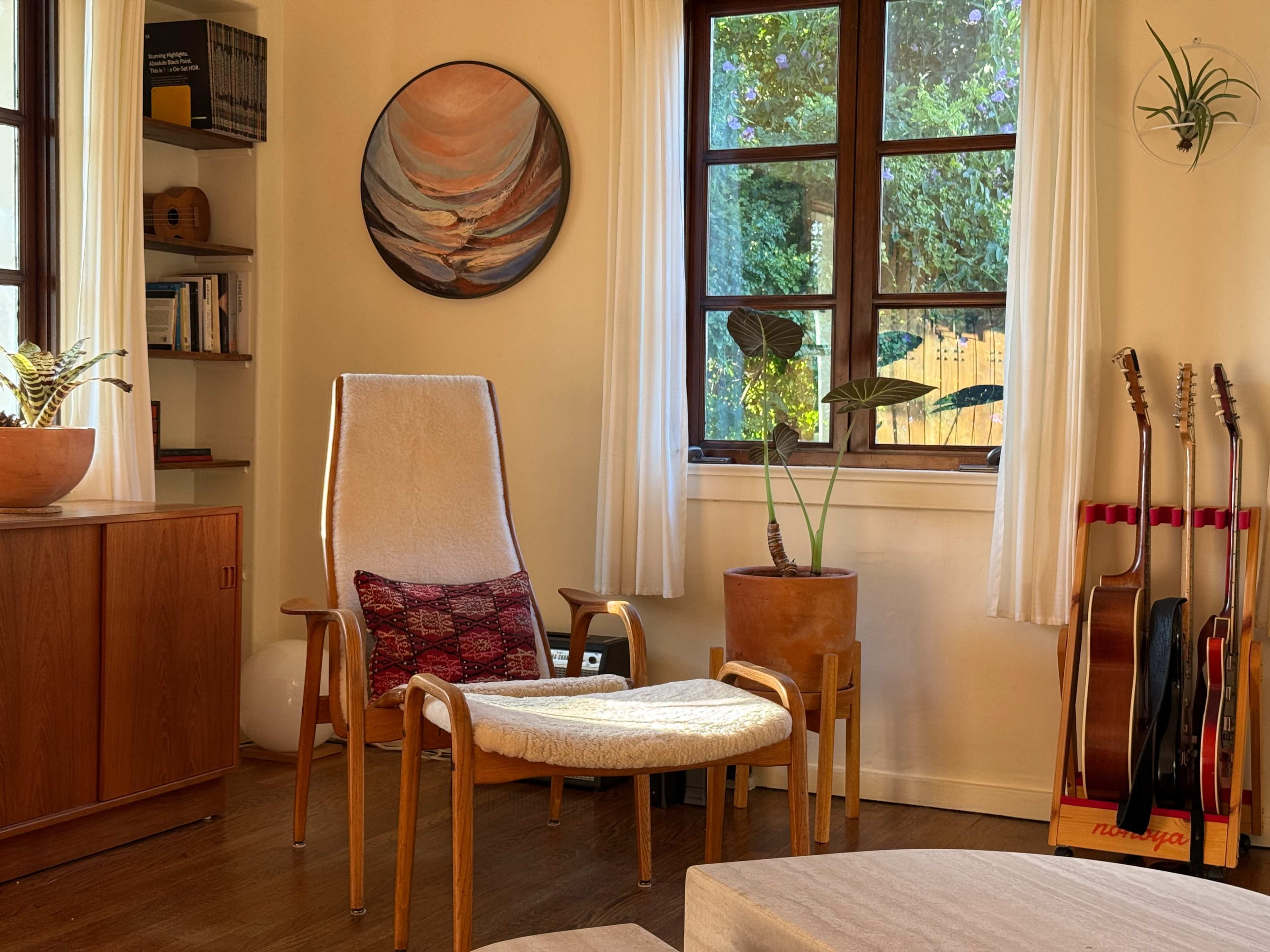 A cozy room features a wooden chair with a white cushion, a round artwork on the wall, a potted plant, and guitars leaning against the wall.