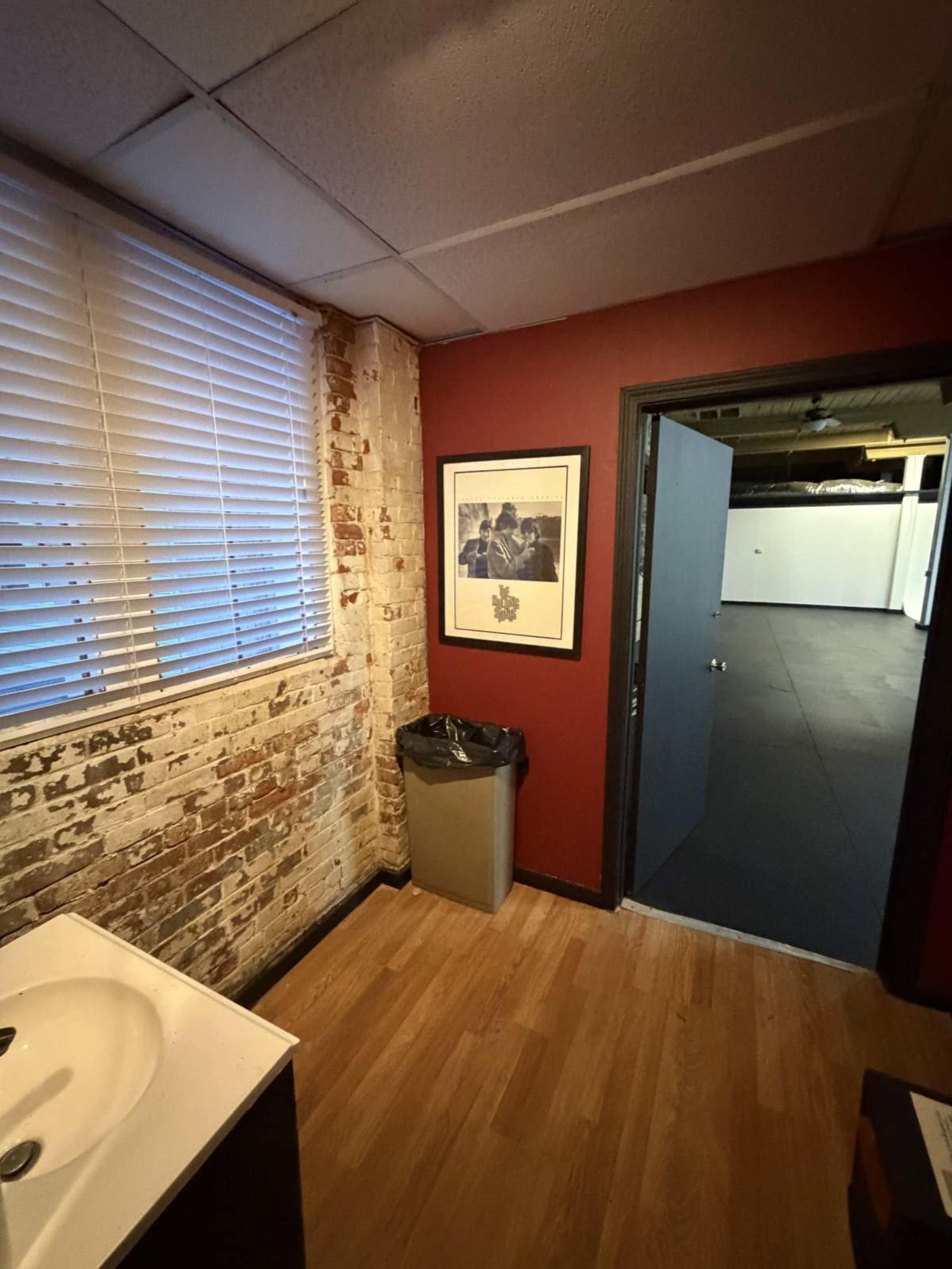 The image shows a small room featuring exposed brick walls, a sink, a framed picture on the wall, a trash can, and a doorway leading to a larger space.