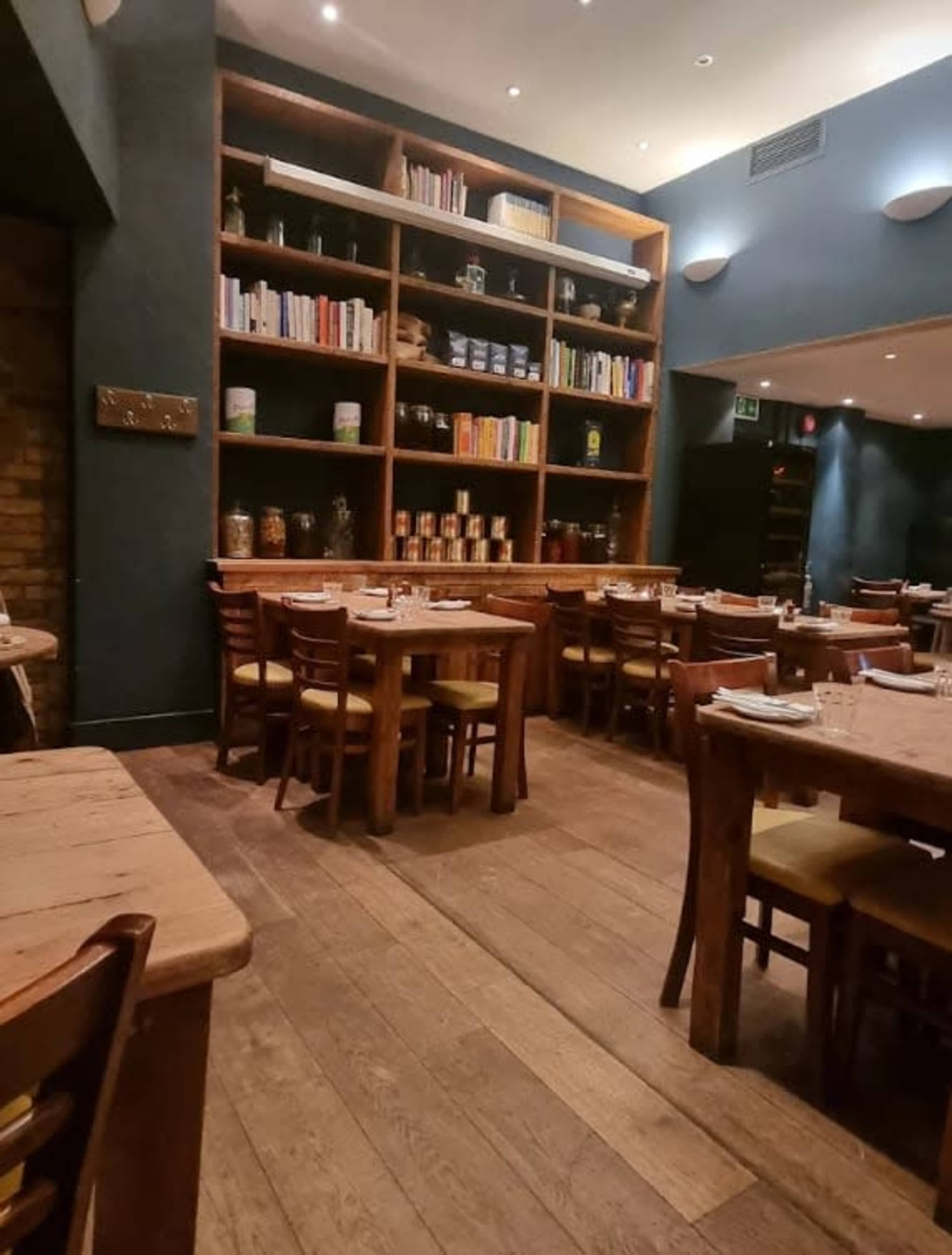 The image shows a cozy restaurant interior with wooden tables, chairs, and shelves filled with books and jars against a dark blue wall.
