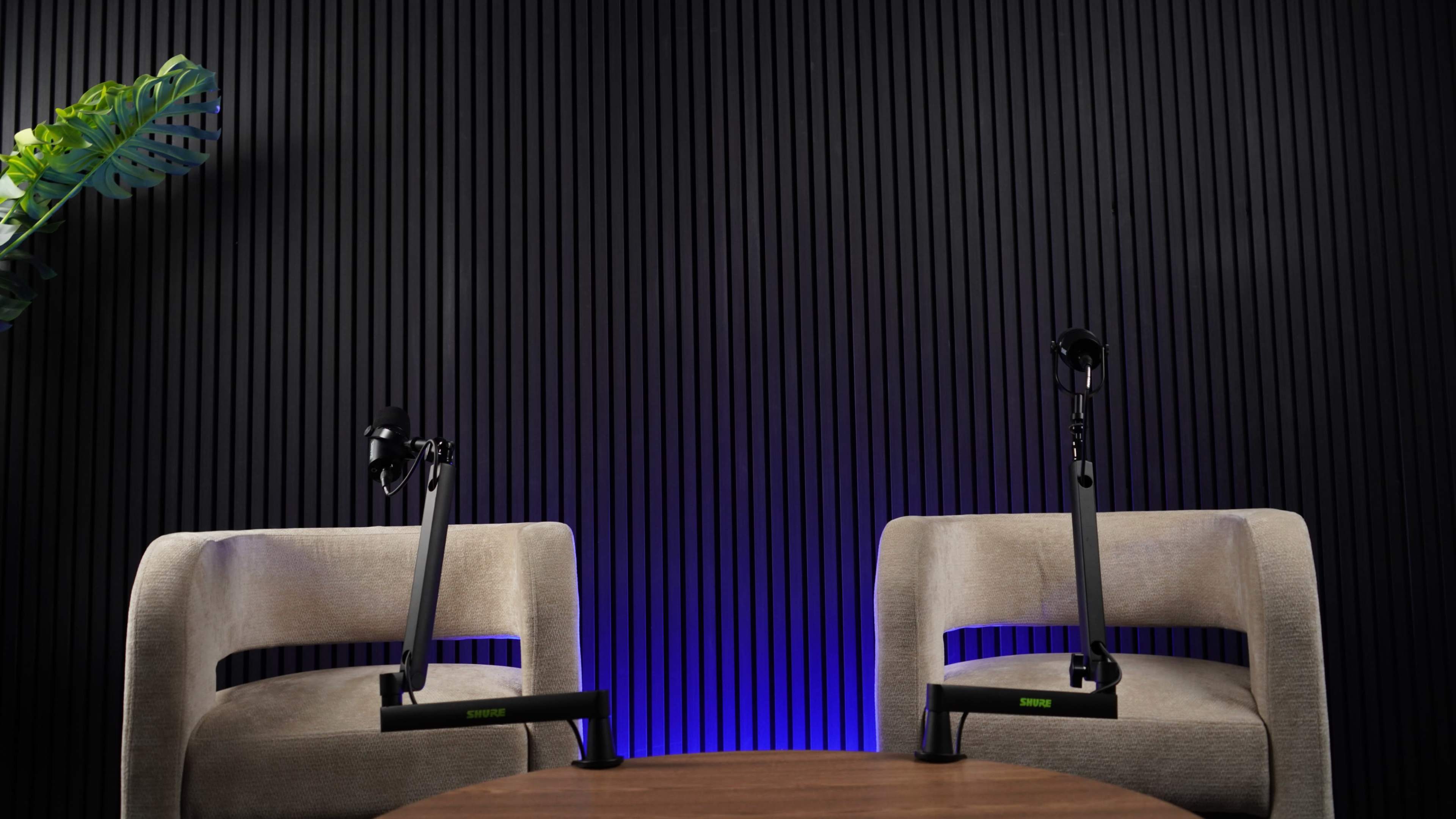 The image shows two empty chairs positioned at a wooden table, each equipped with microphone stands, against a textured black wall with blue lighting.