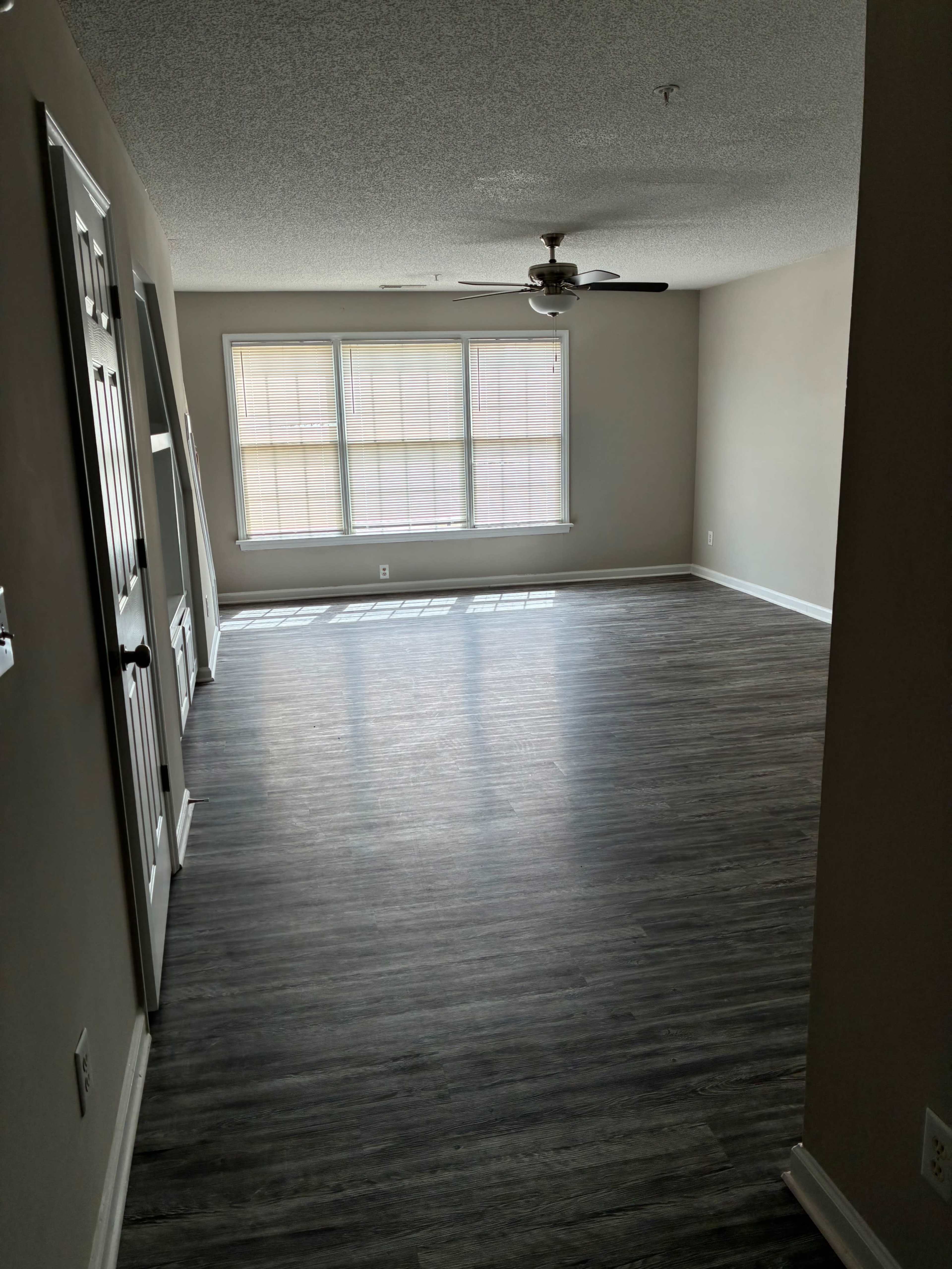 The image shows an empty room with a ceiling fan and large windows, featuring a hardwood floor and light-colored walls.
