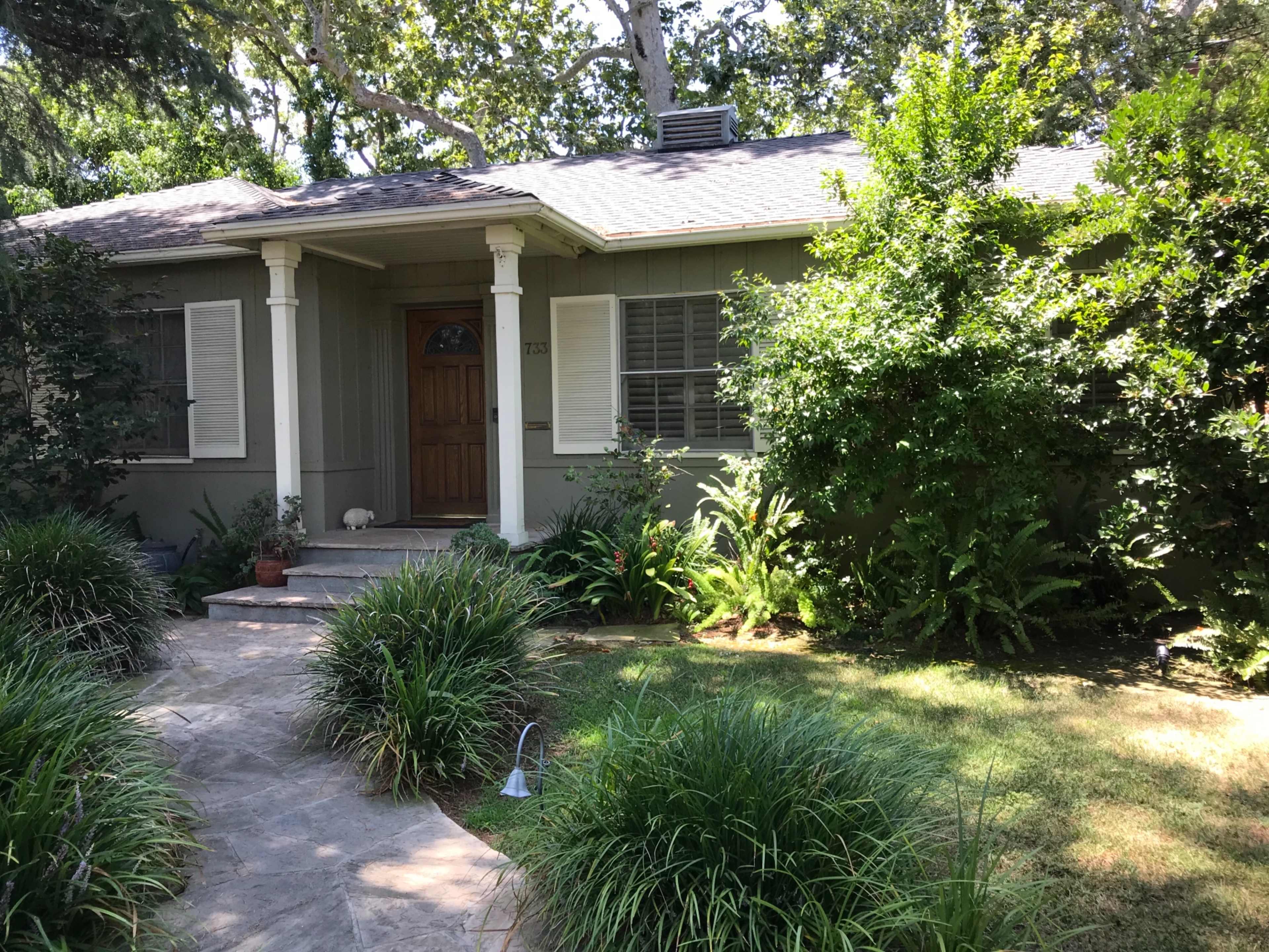 A single-story house with a wooden front door is surrounded by a landscaped garden featuring shrubs and grass.