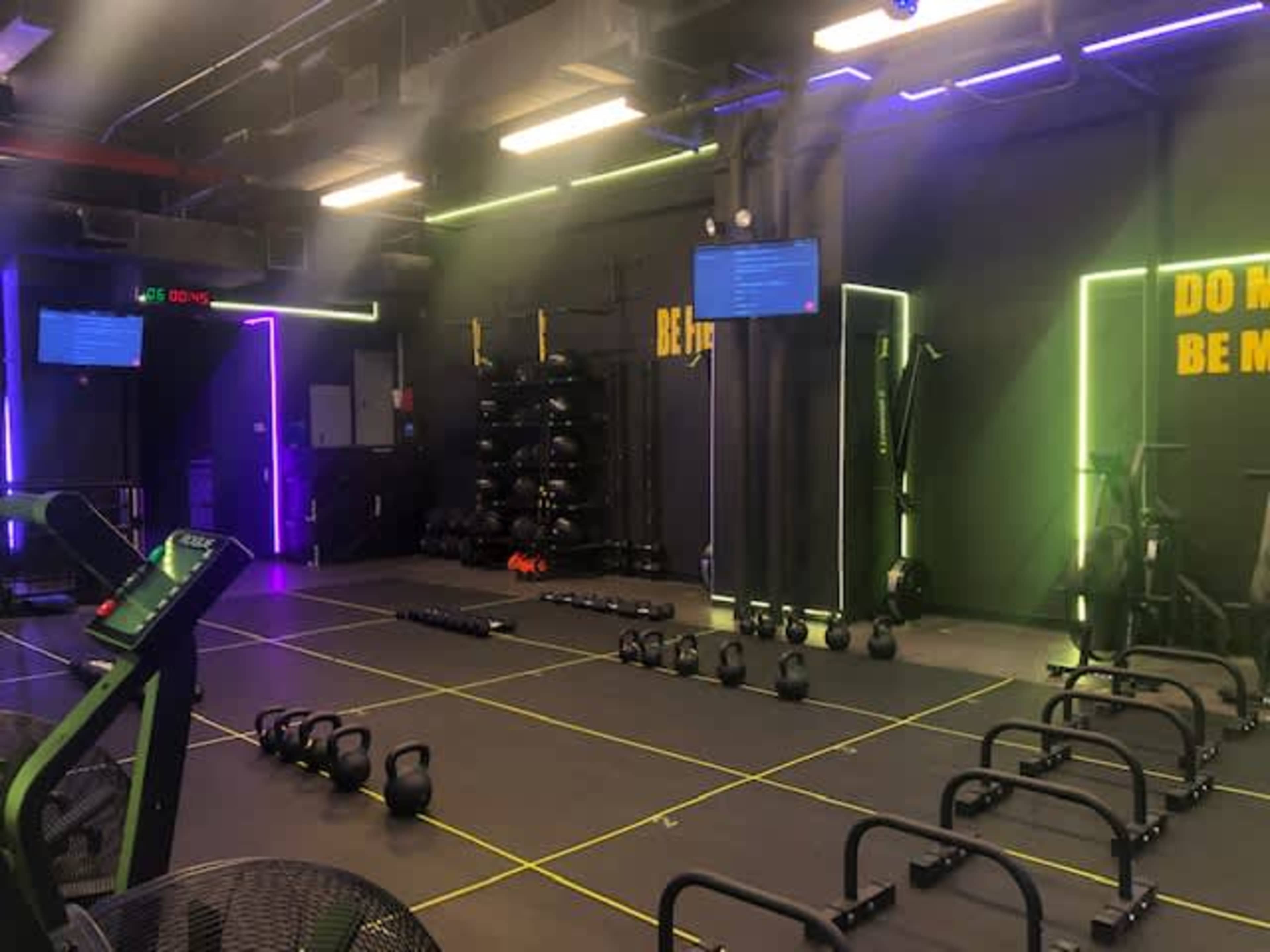 The image shows a fitness studio equipped with workout stations, kettlebells, and wall-mounted screens.
