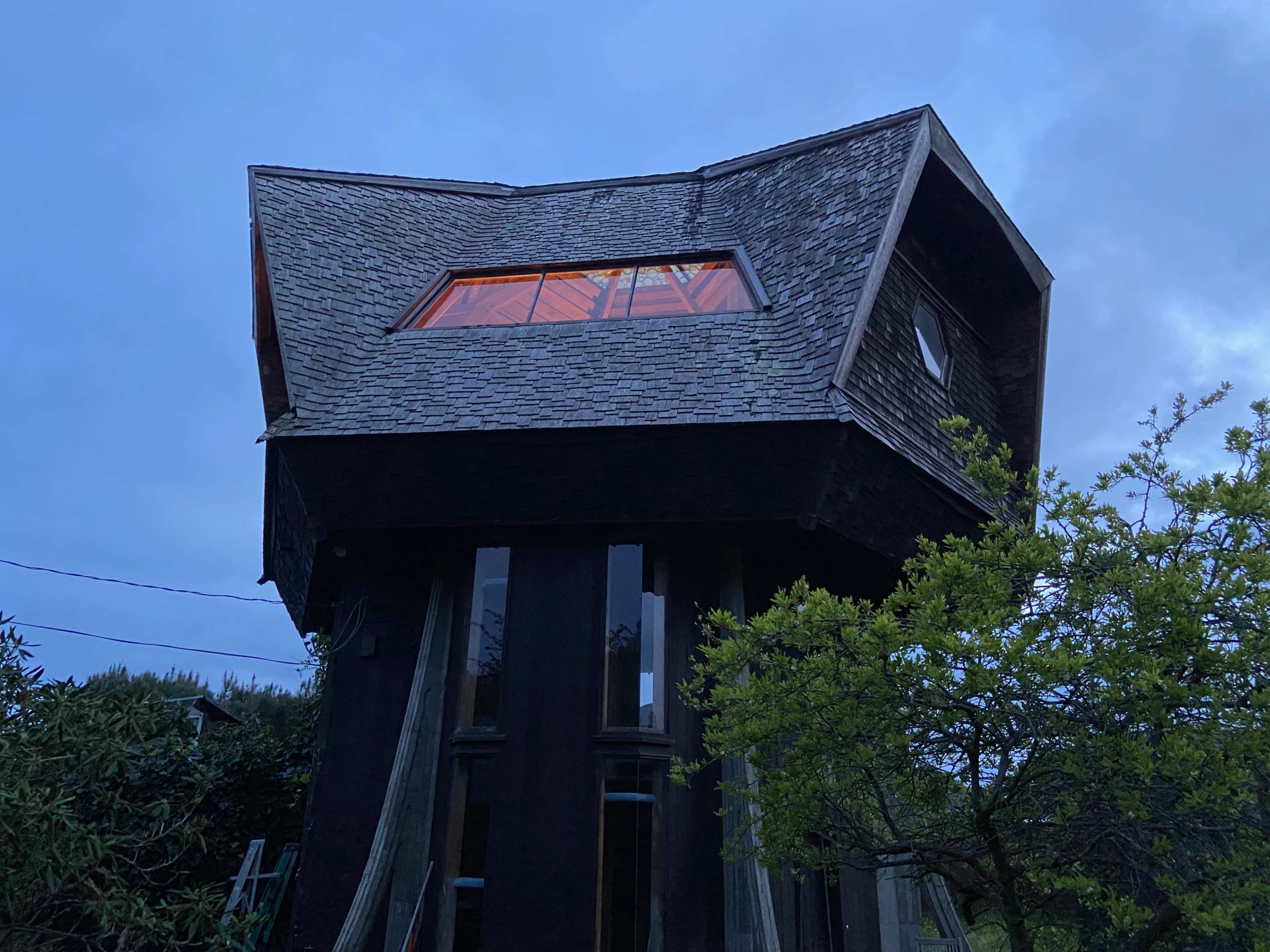 A unique, angled wooden structure stands on stilts amidst a cloudy sky, showcasing large windows that reflect an orange glow.