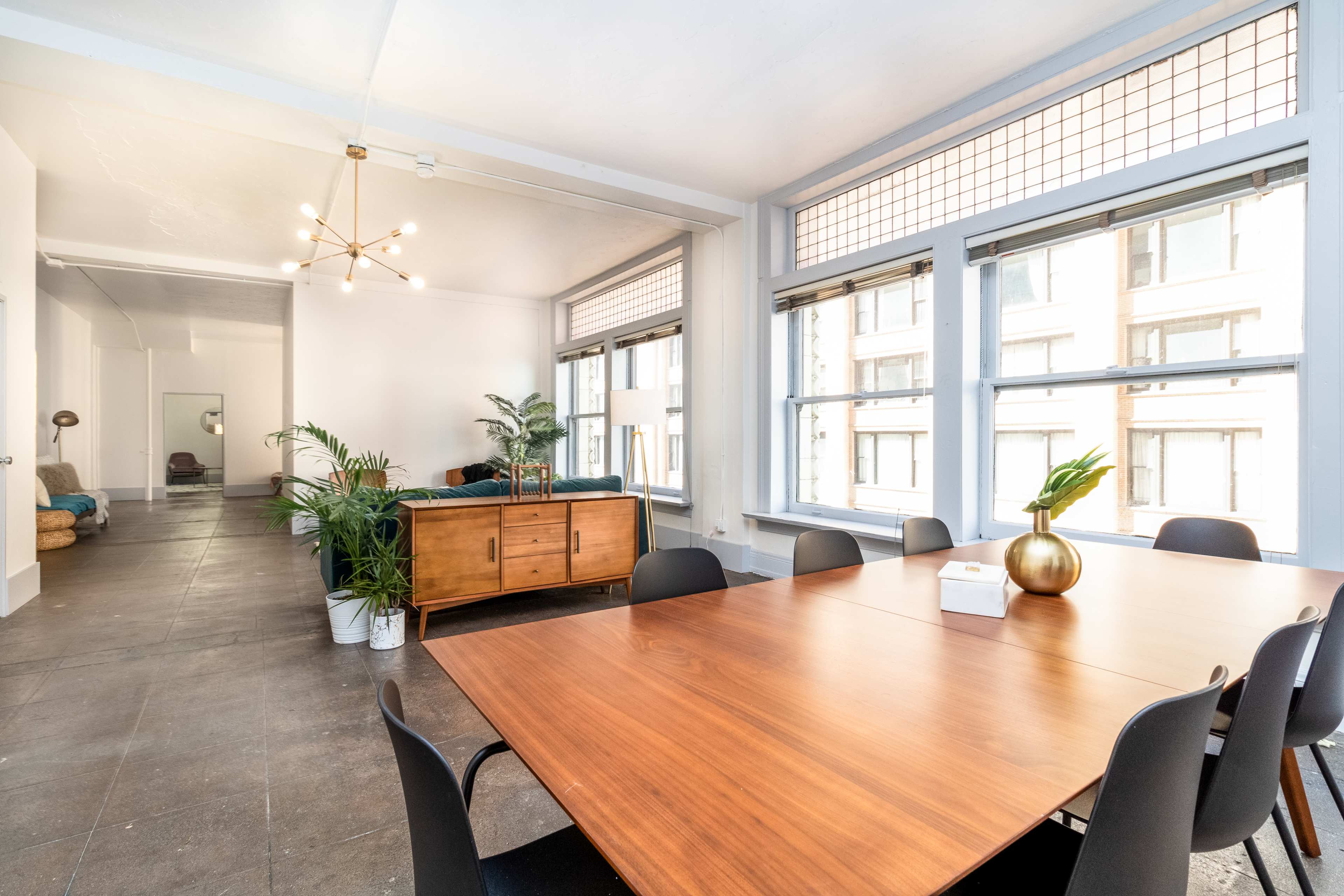 The image shows a spacious room with a large wooden table surrounded by chairs, windows allowing natural light, and a stylistic light fixture overhead.
