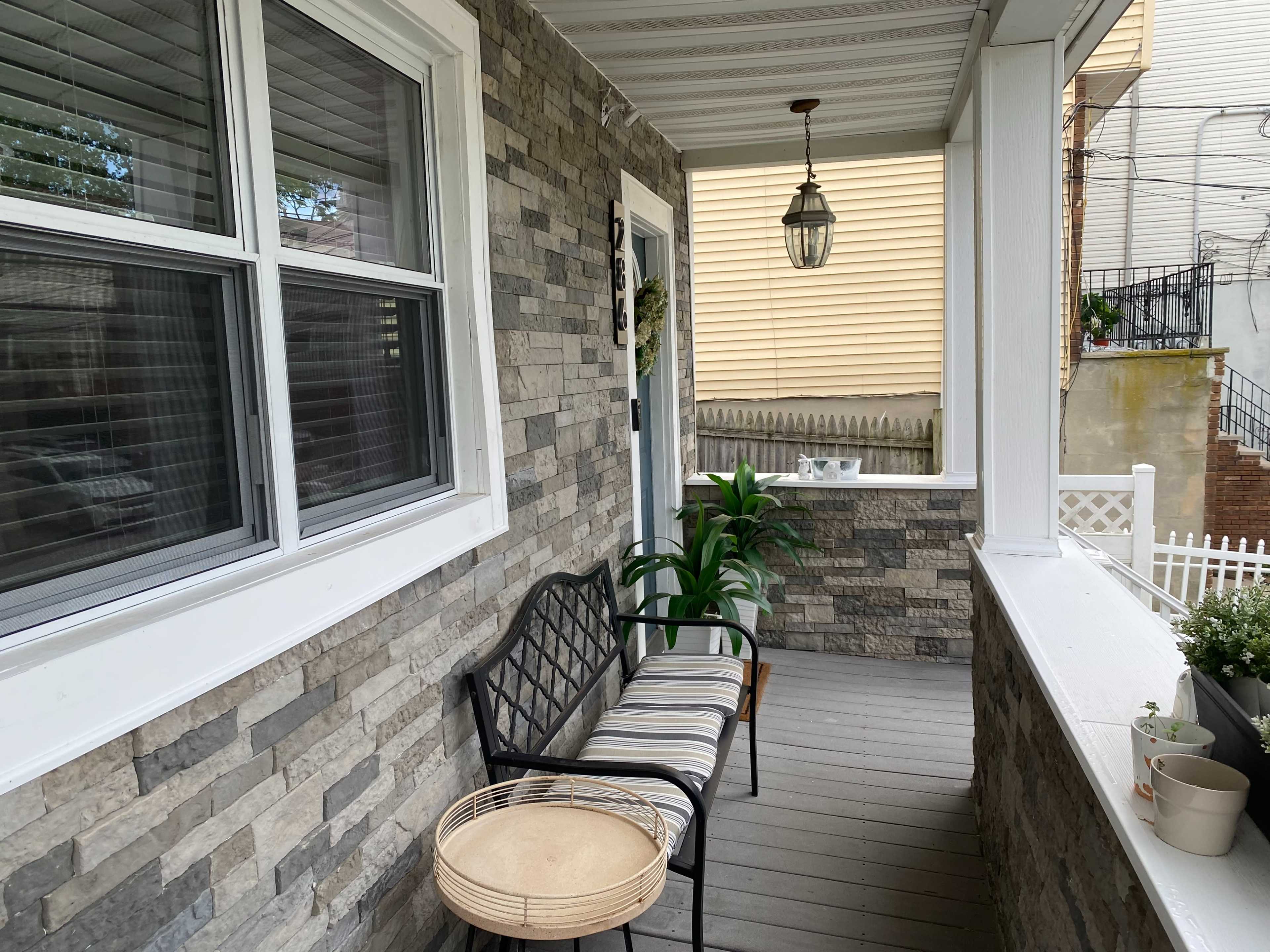 The image shows a porch with stone walls, a bench with striped cushions, a round table, and a green plant.